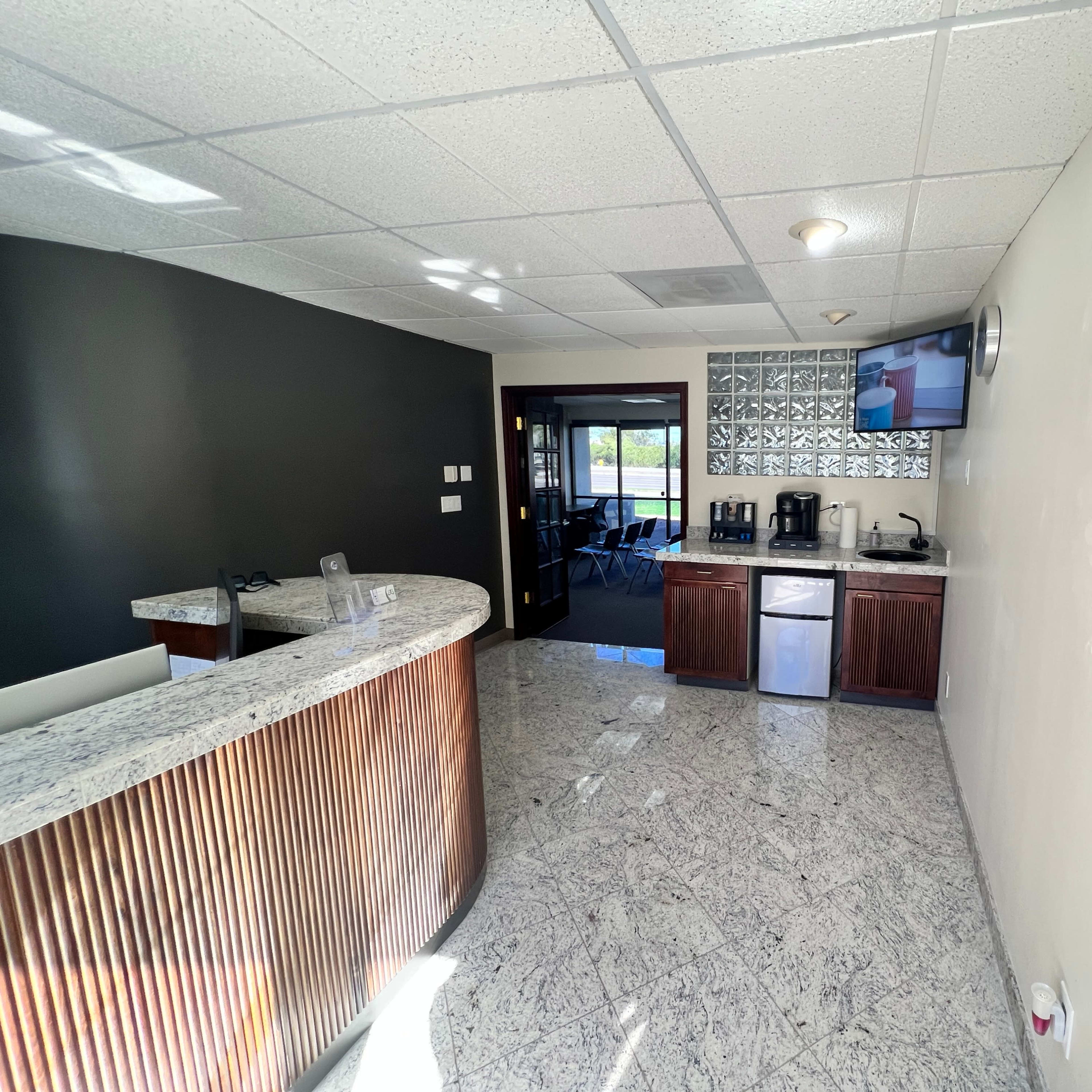 A modern reception area with a curved front desk, a coffee station, a refrigerator, and a large window overlooking a meeting space.