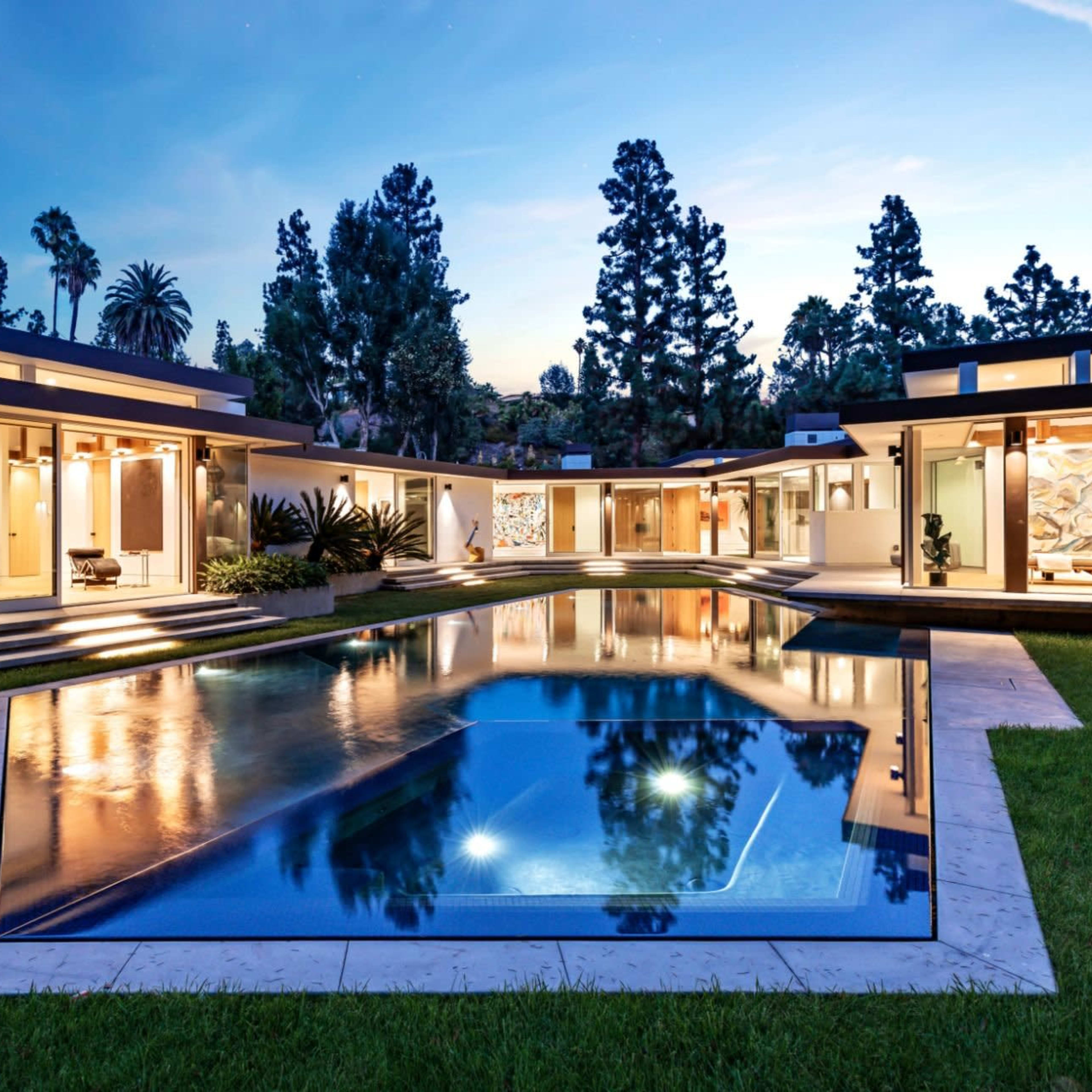 A modern house with an expansive pool is surrounded by landscaped gardens and tall trees under a twilight sky.