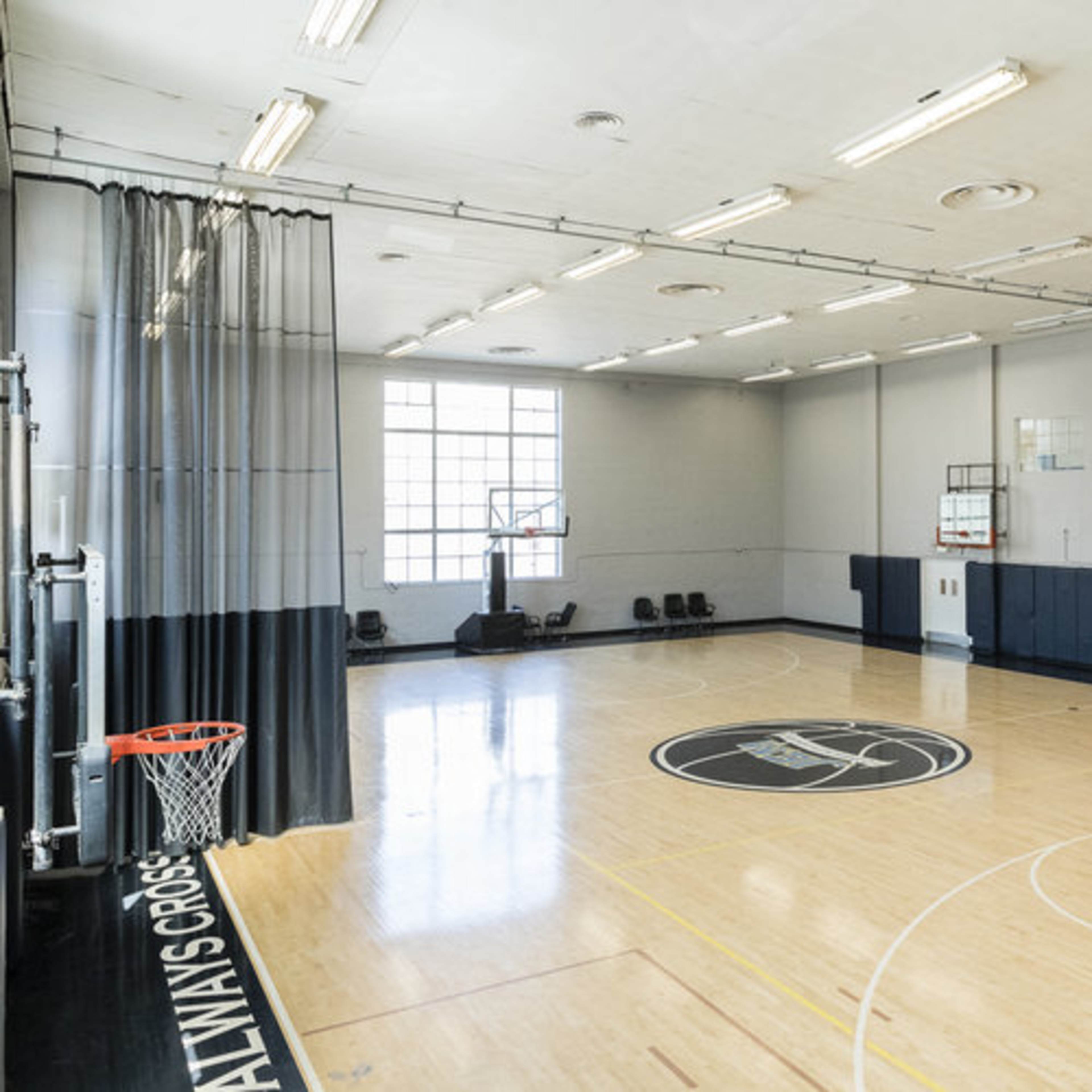 A spacious indoor basketball court with a polished wooden floor, basketball hoops on both ends, and a curtain dividing the court area.