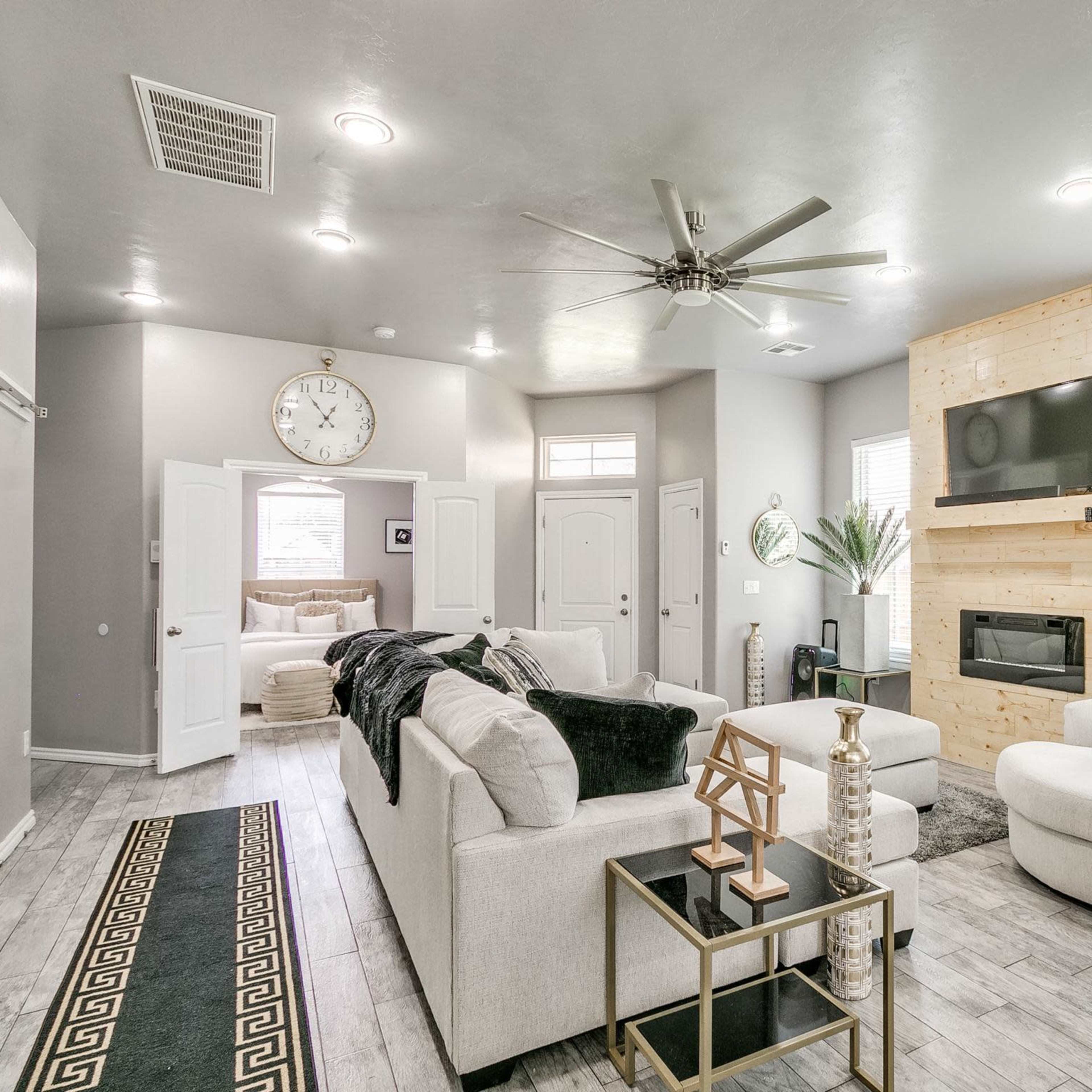 A modern living room with a light color palette, featuring a sectional sofa, two armchairs, a coffee table, a mounted television, and a decorative fireplace.