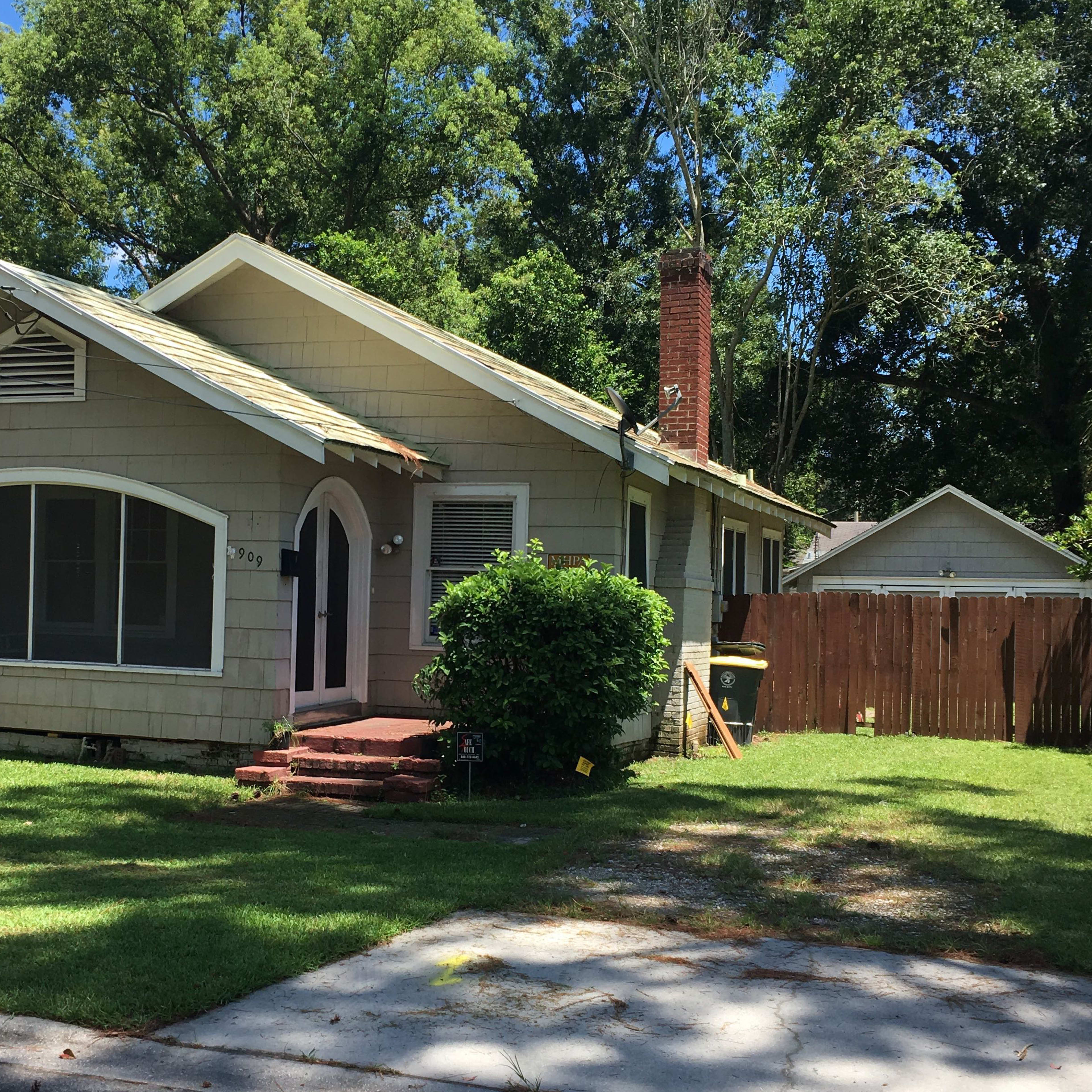 A single-story, light-colored house with a sloped roof and a front porch is surrounded by green grass and trees.