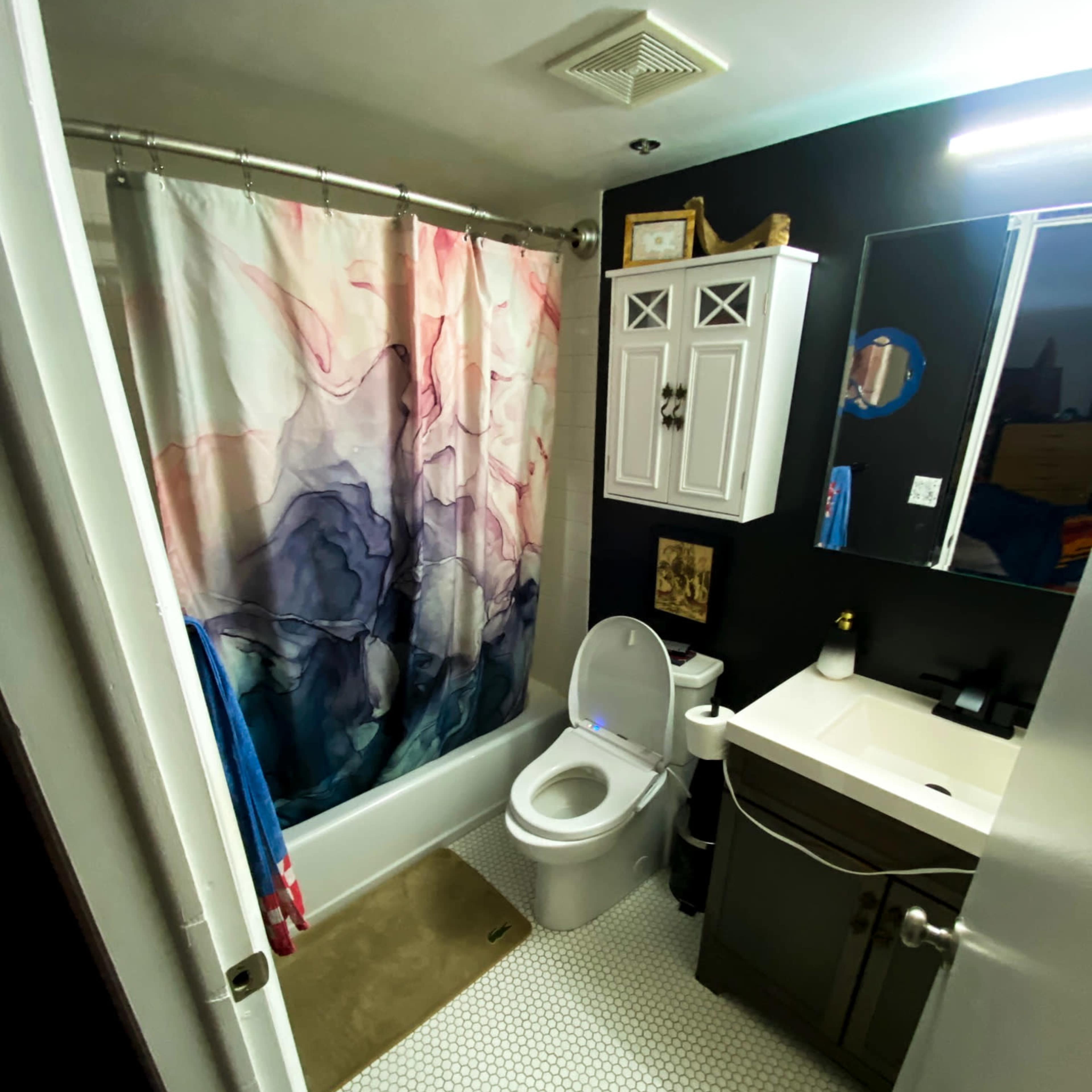 A compact bathroom featuring a shower with a colorful curtain, a toilet, a sink with a cabinet, and hexagonal tile flooring.