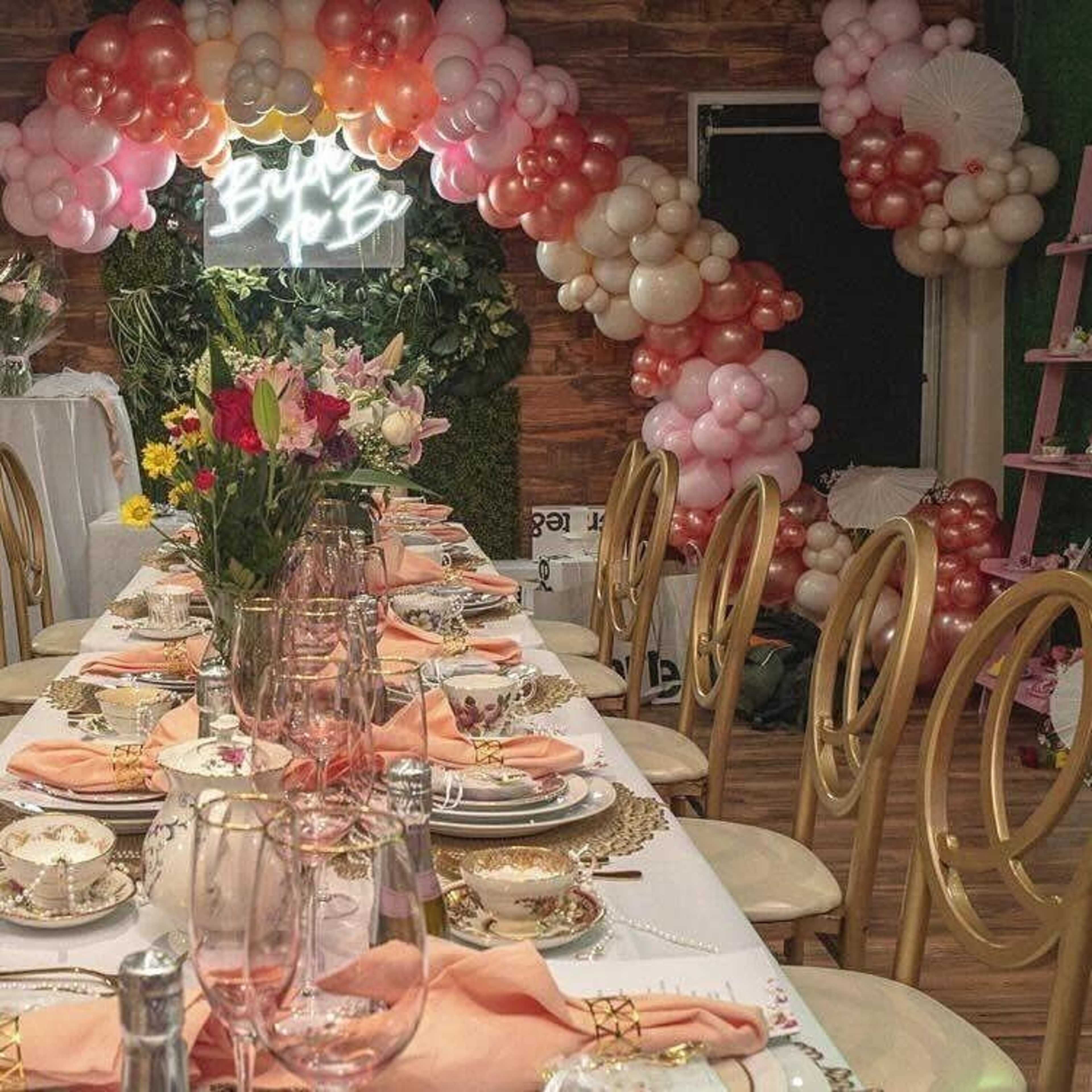 A long dining table is elegantly set with floral centerpieces, place settings, and a backdrop of pink and white balloon decorations.