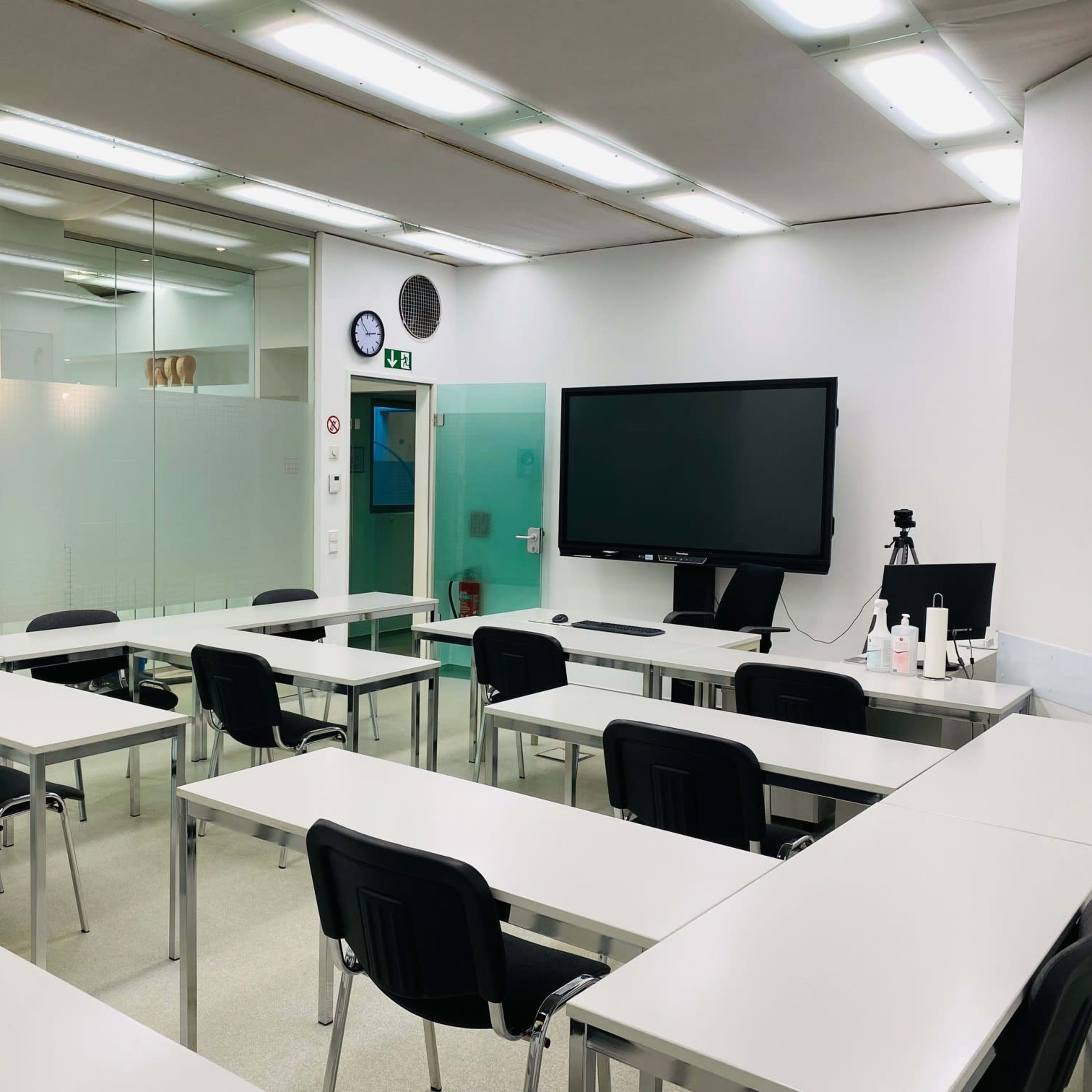 A classroom is set up with several tables and chairs facing a large screen, under bright fluorescent lighting.