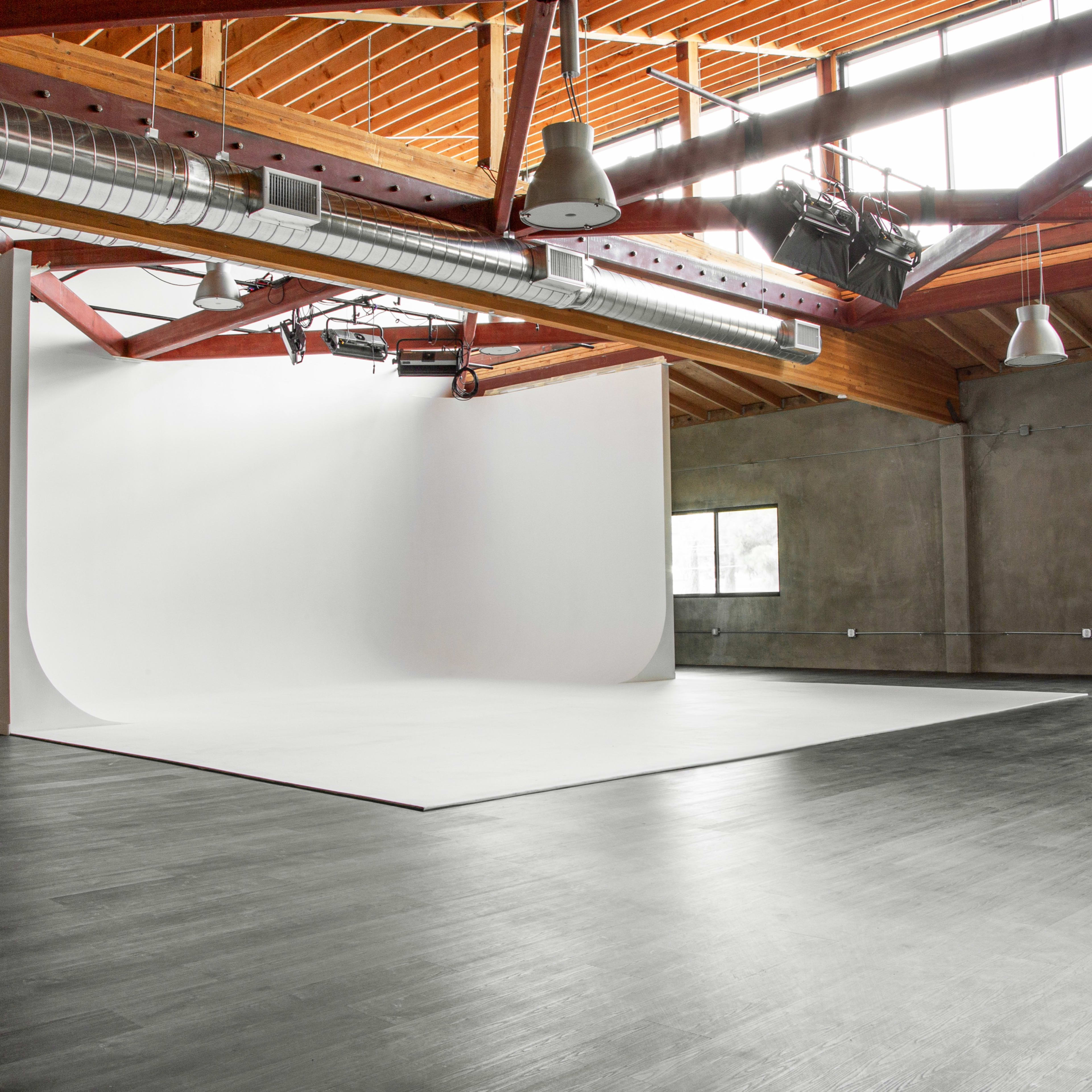 A spacious photography studio with a seamless white backdrop, high wooden ceilings, and large windows allowing natural light.