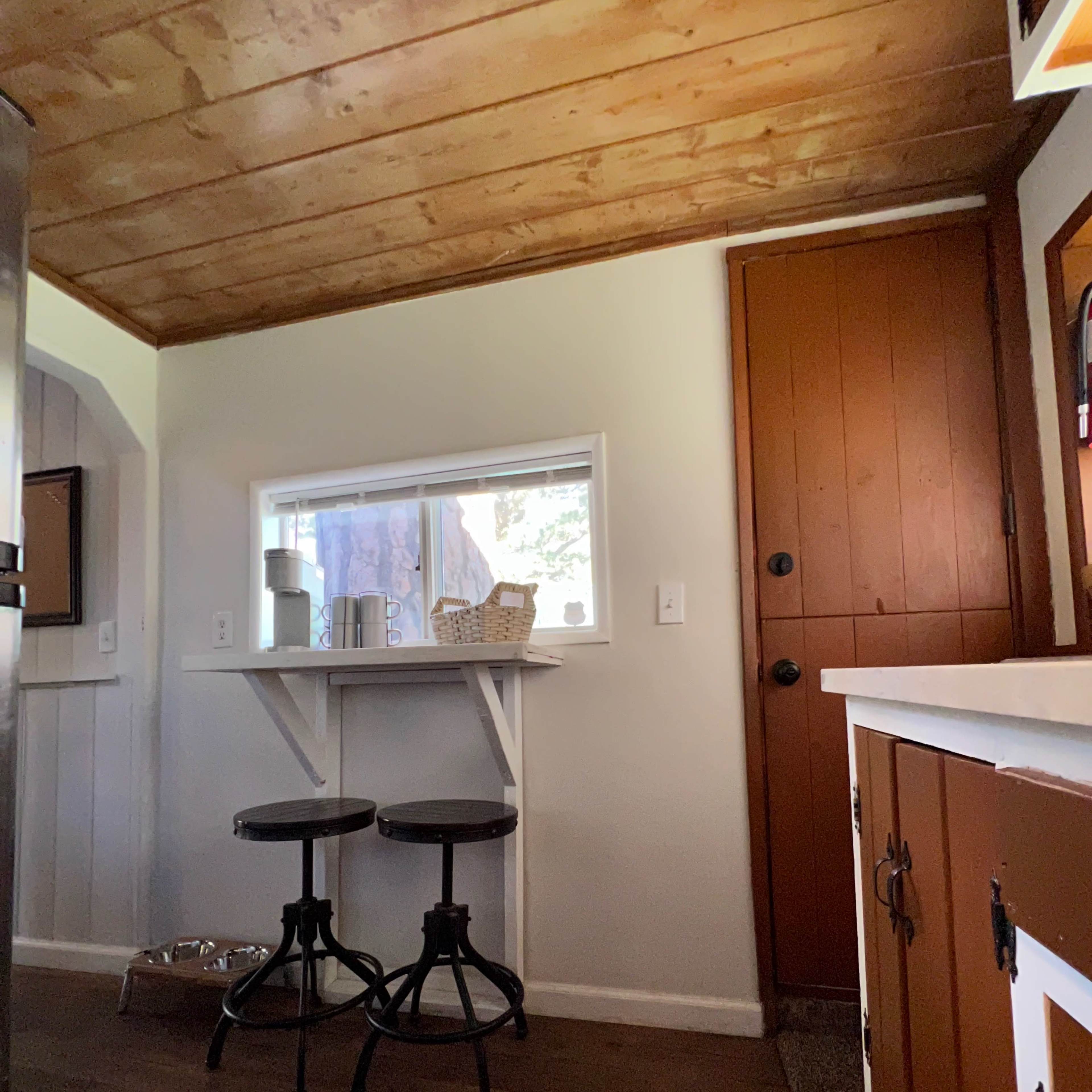 A kitchen space with a wooden ceiling, a small table with two black stools, a door painted in a reddish-brown color, and a window that allows natural light to enter.