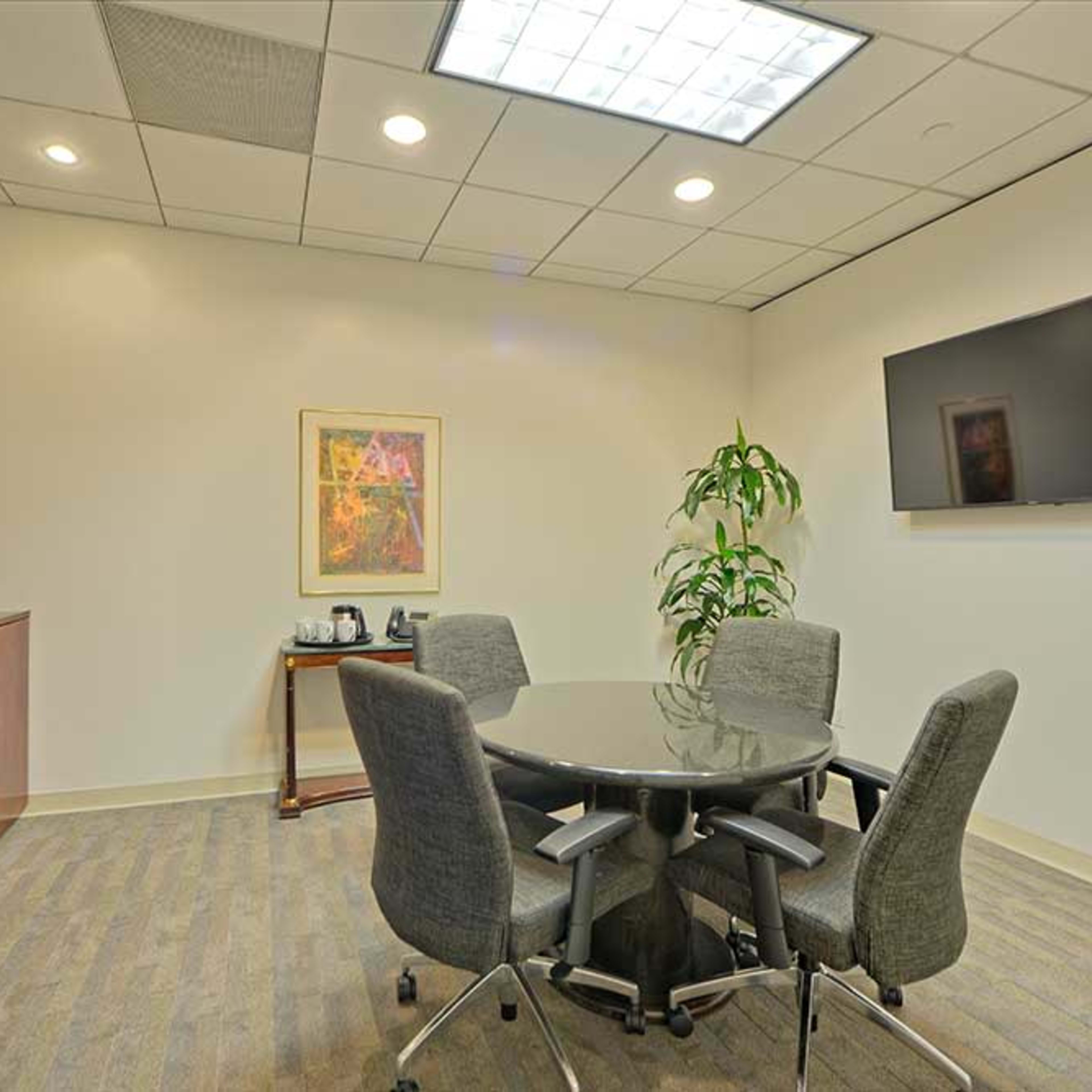 A meeting room features a round table surrounded by four chairs, a wall-mounted television, and a tall plant in the corner.