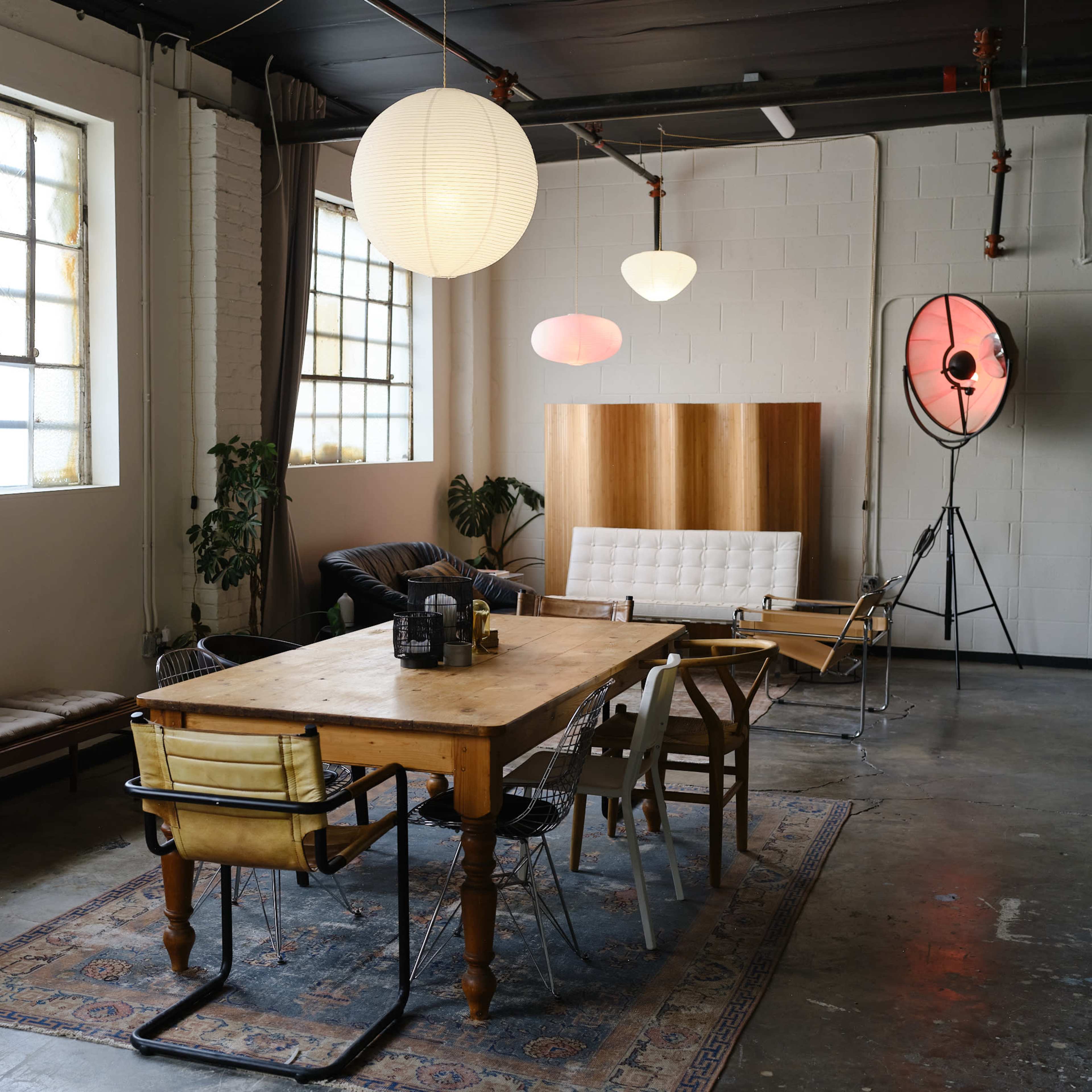 A spacious interior featuring a wooden dining table surrounded by various chairs, large windows, and soft lighting fixtures.