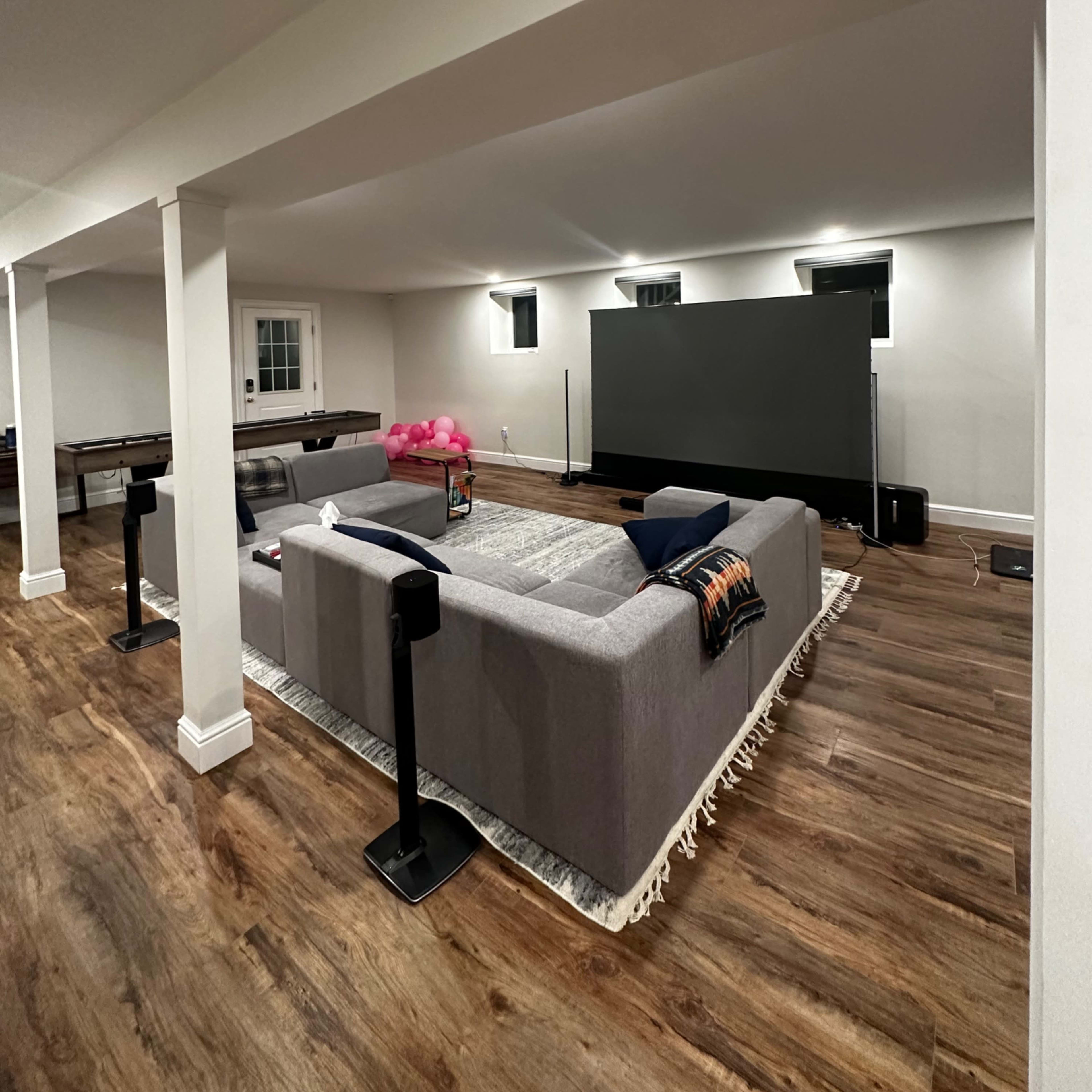 A spacious basement living area features a large gray sectional sofa facing a wall-mounted television, with wooden flooring and lit windows.