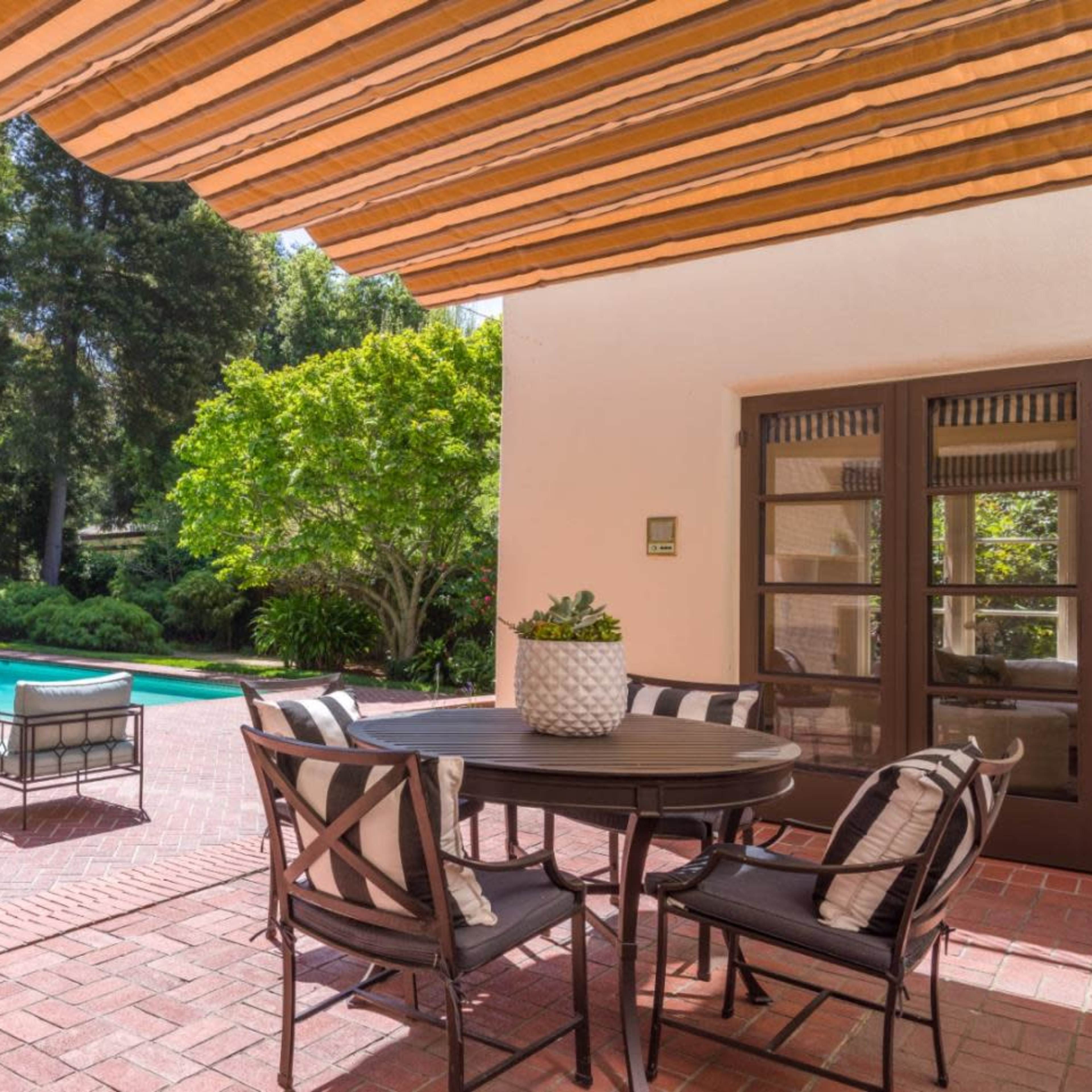 A brick patio with a dining table and chairs overlooks a pool surrounded by trees and landscaped greenery.