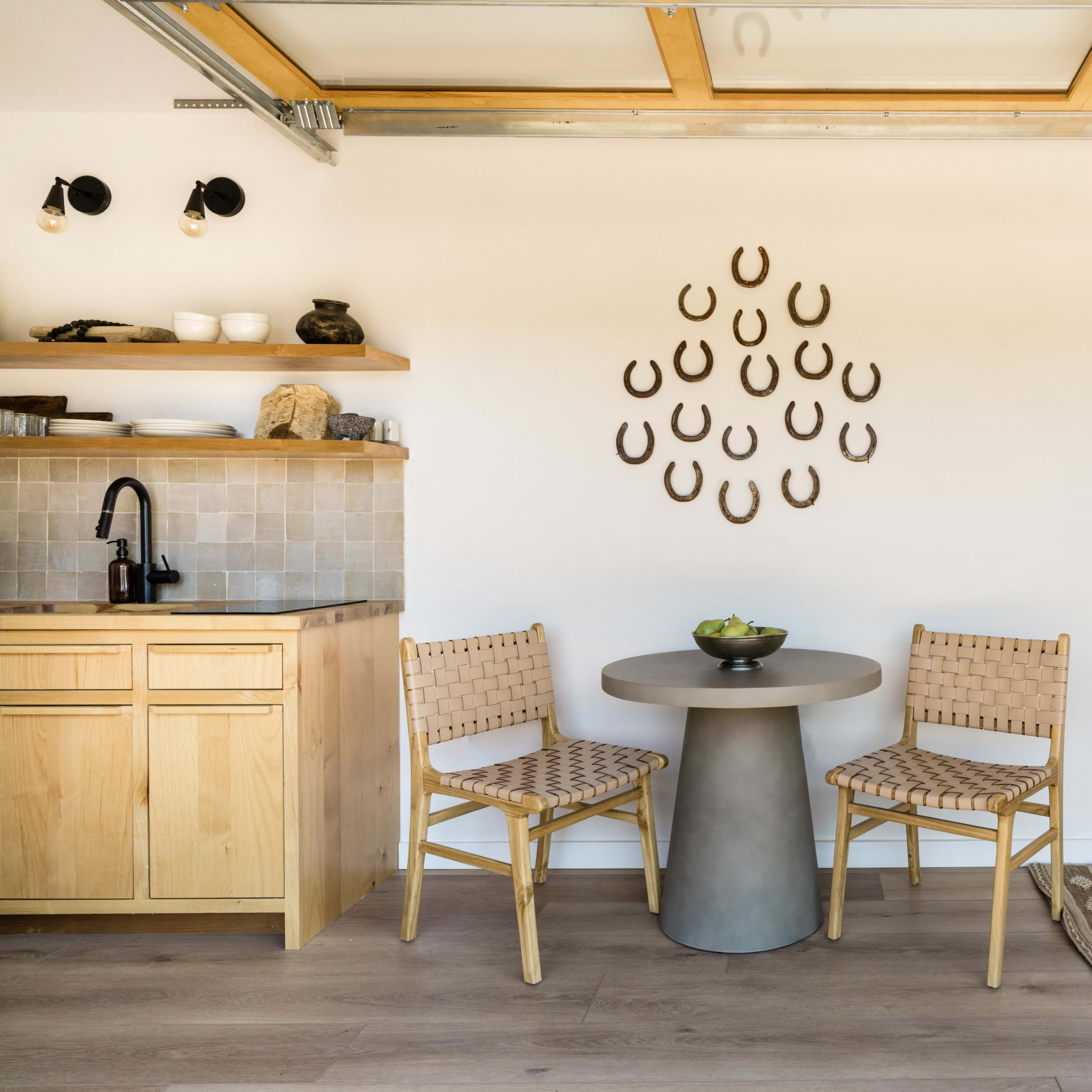 A minimalist kitchen area features wooden cabinets, a round table with two woven chairs, and a cozy seating space, accented by a wall display of horseshoes.