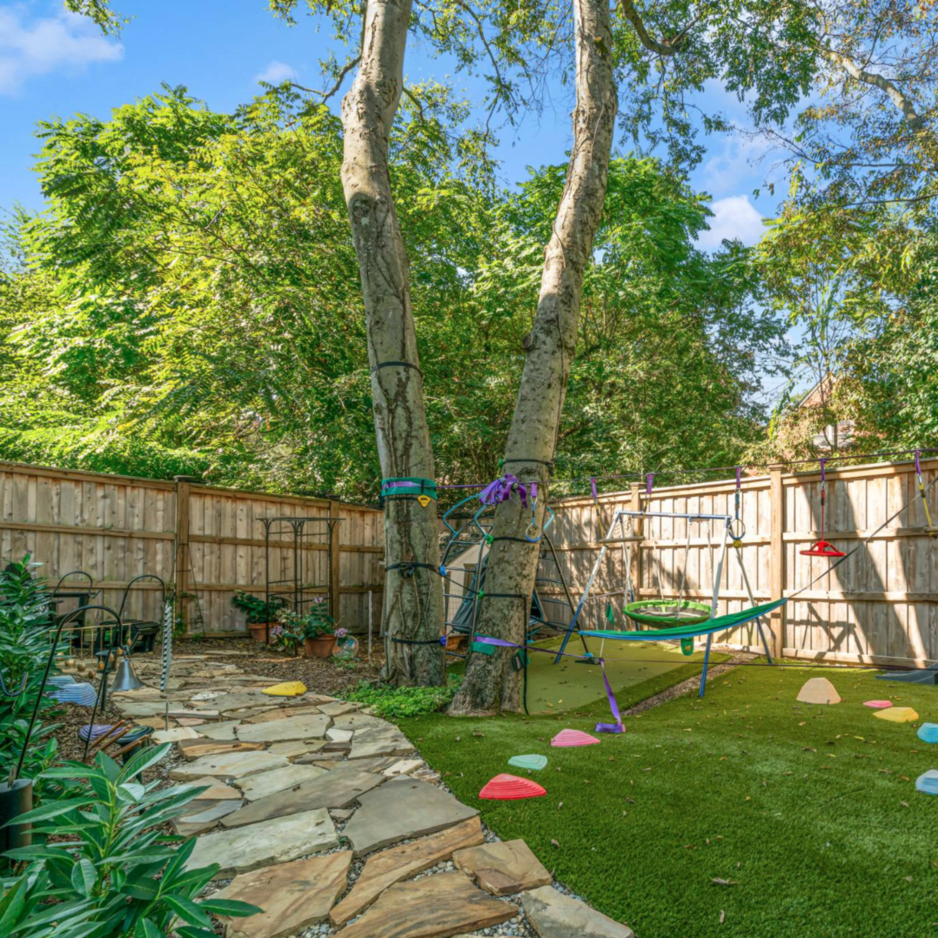 A backyard with a stone pathway, a play structure, and colorful stepping stones on a grassy area surrounded by wooden fencing and trees.