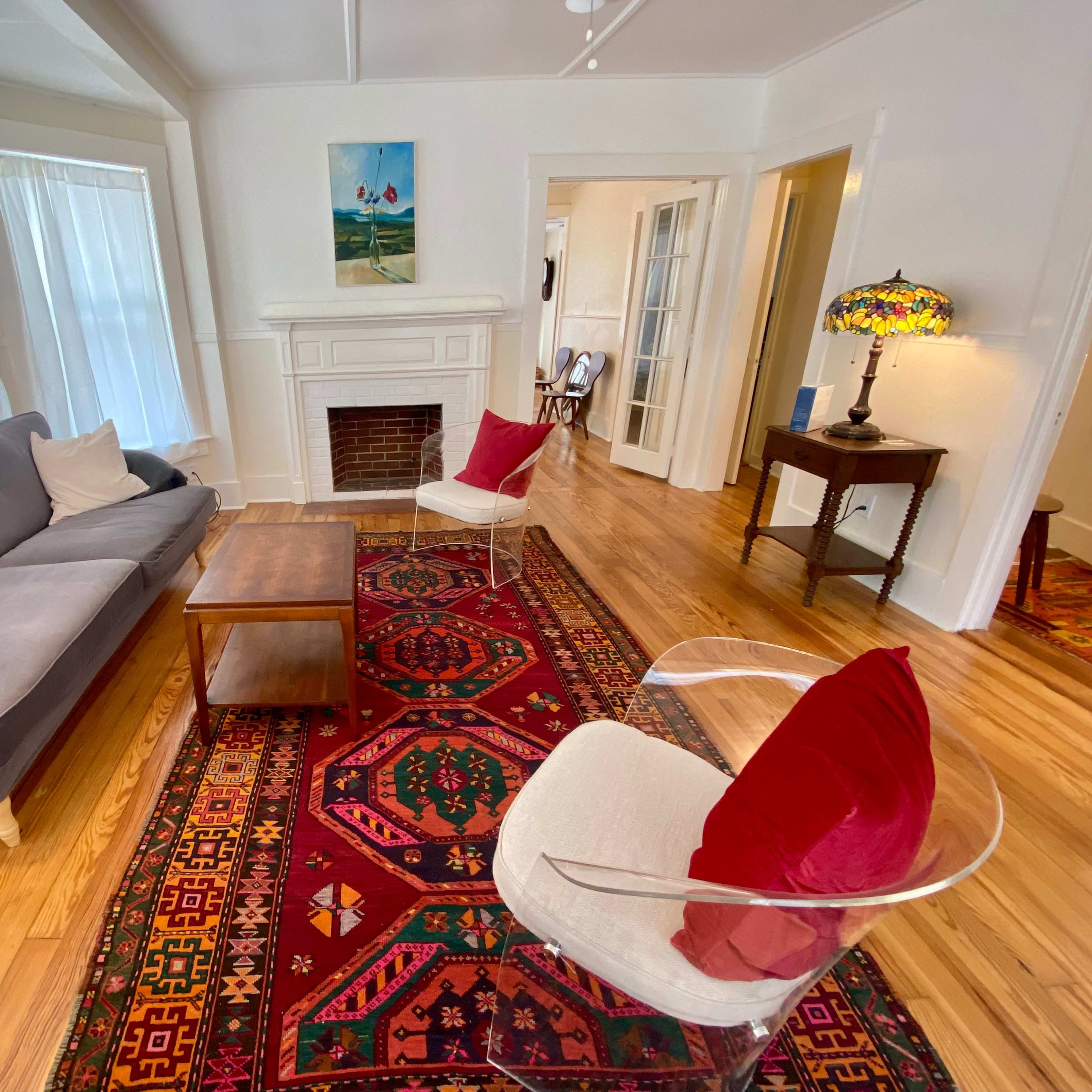 The living room features a gray sofa, two white chairs with red cushions, a wooden coffee table, and a colorful rug, along with a fireplace and artwork on the walls.