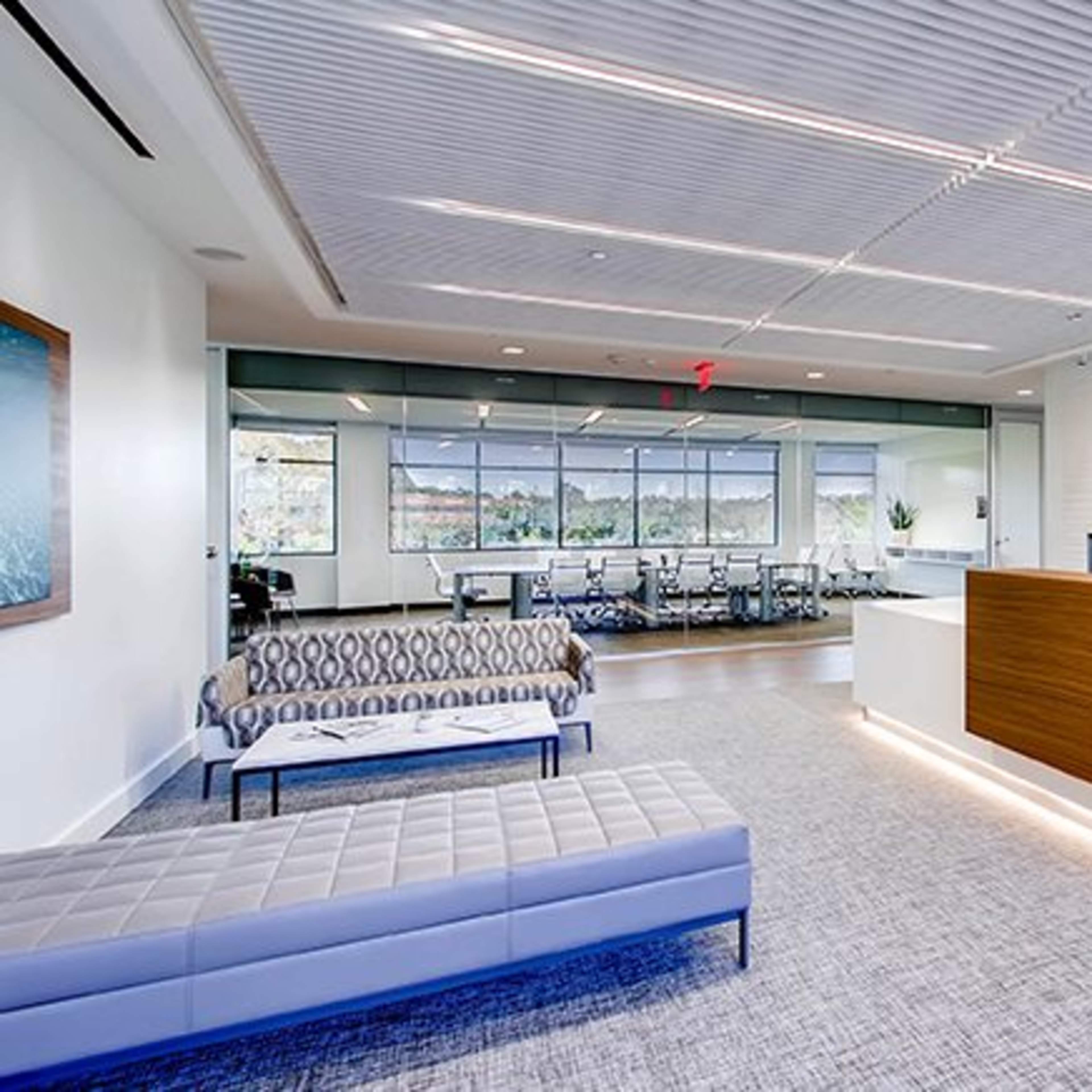 A modern office reception area featuring a wooden front desk, seating arrangements with sofas, and a large conference room visible in the background.