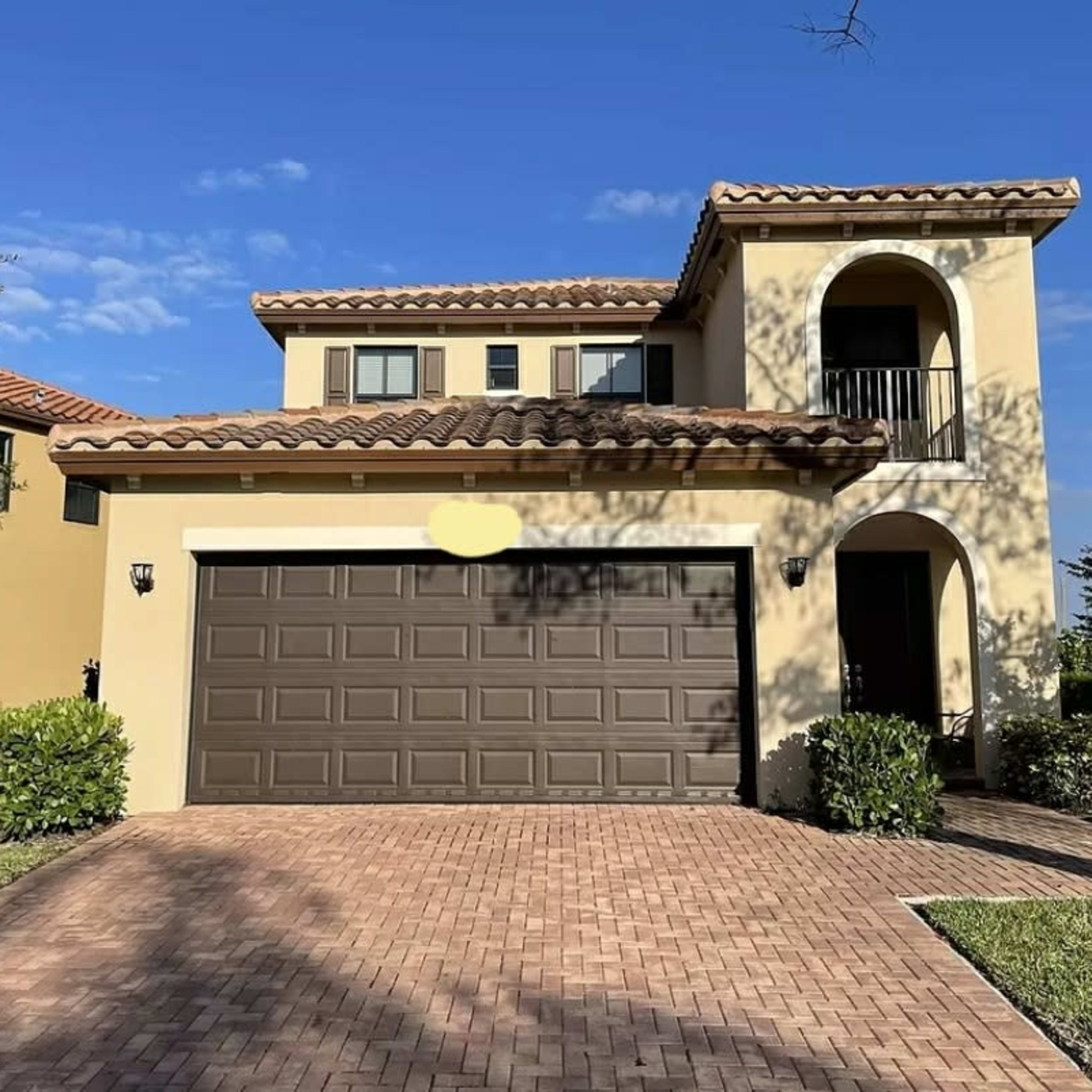 A two-story house with a brown garage door and a brick driveway is set against a blue sky.