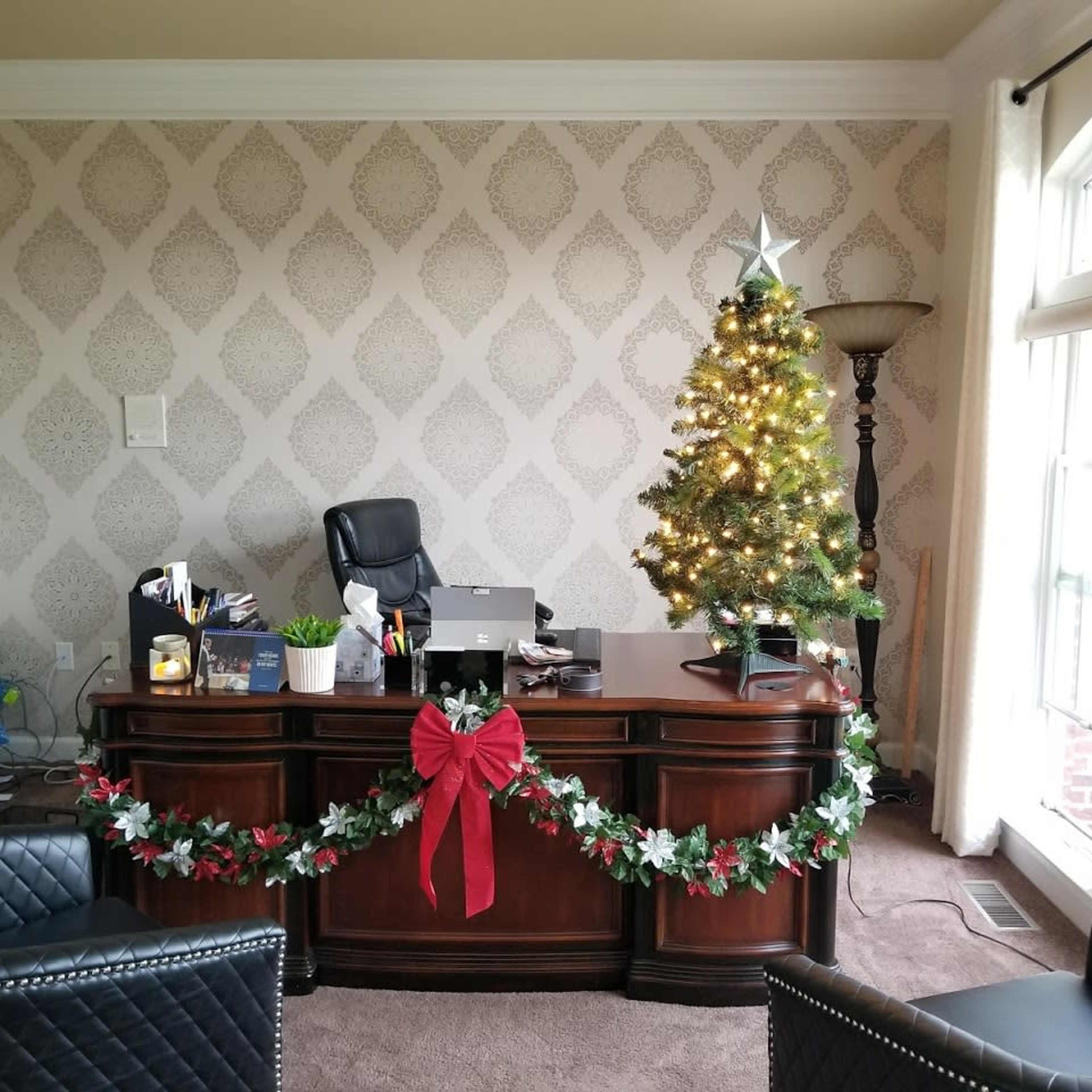 A neatly arranged office space featuring a wooden desk with a computer, an office chair, and a small Christmas tree decorated with lights, placed beside the desk and adorned with a garland on the front.