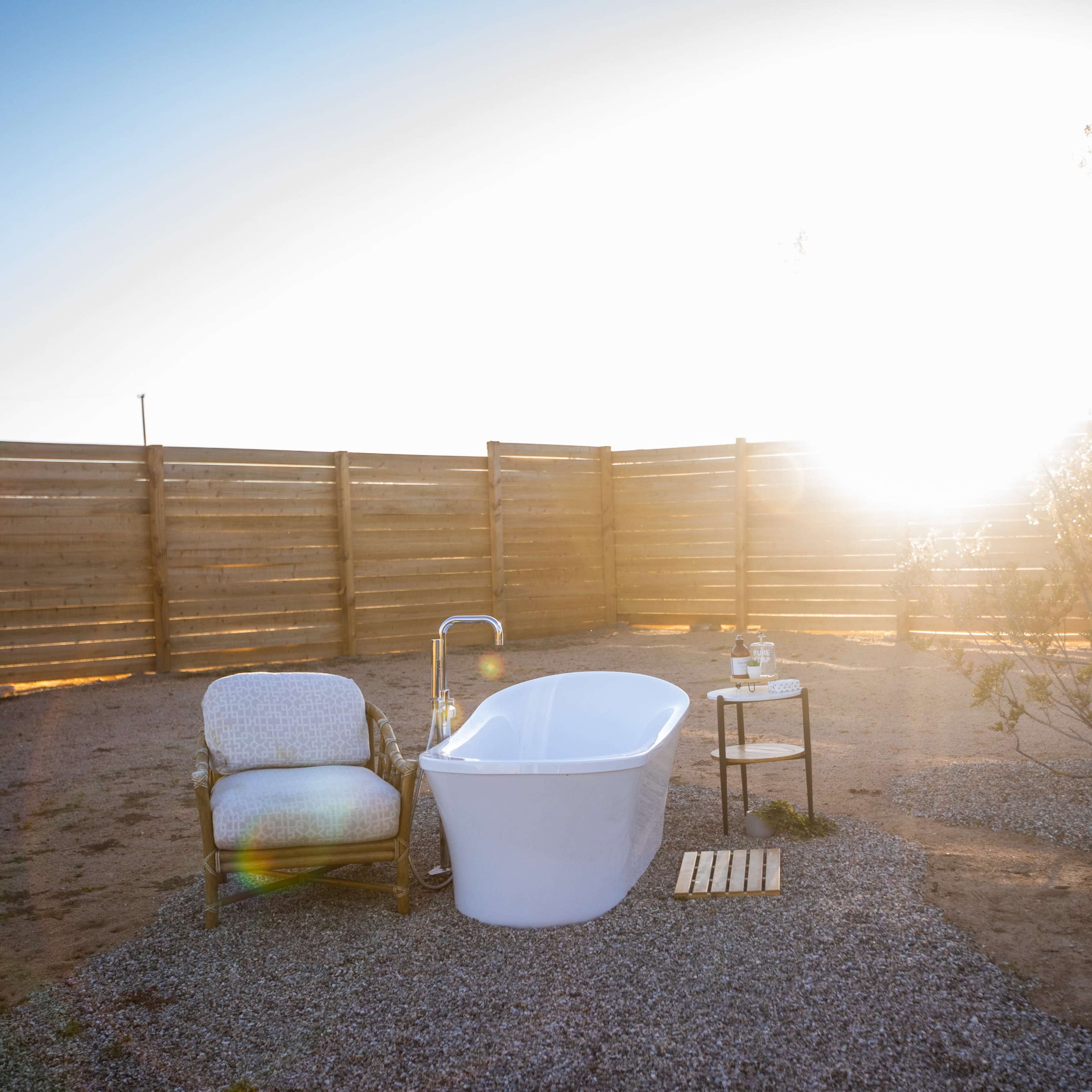 A white freestanding bathtub and a chair are set on a gravel area surrounded by a wooden fence, with the sun shining in the background.