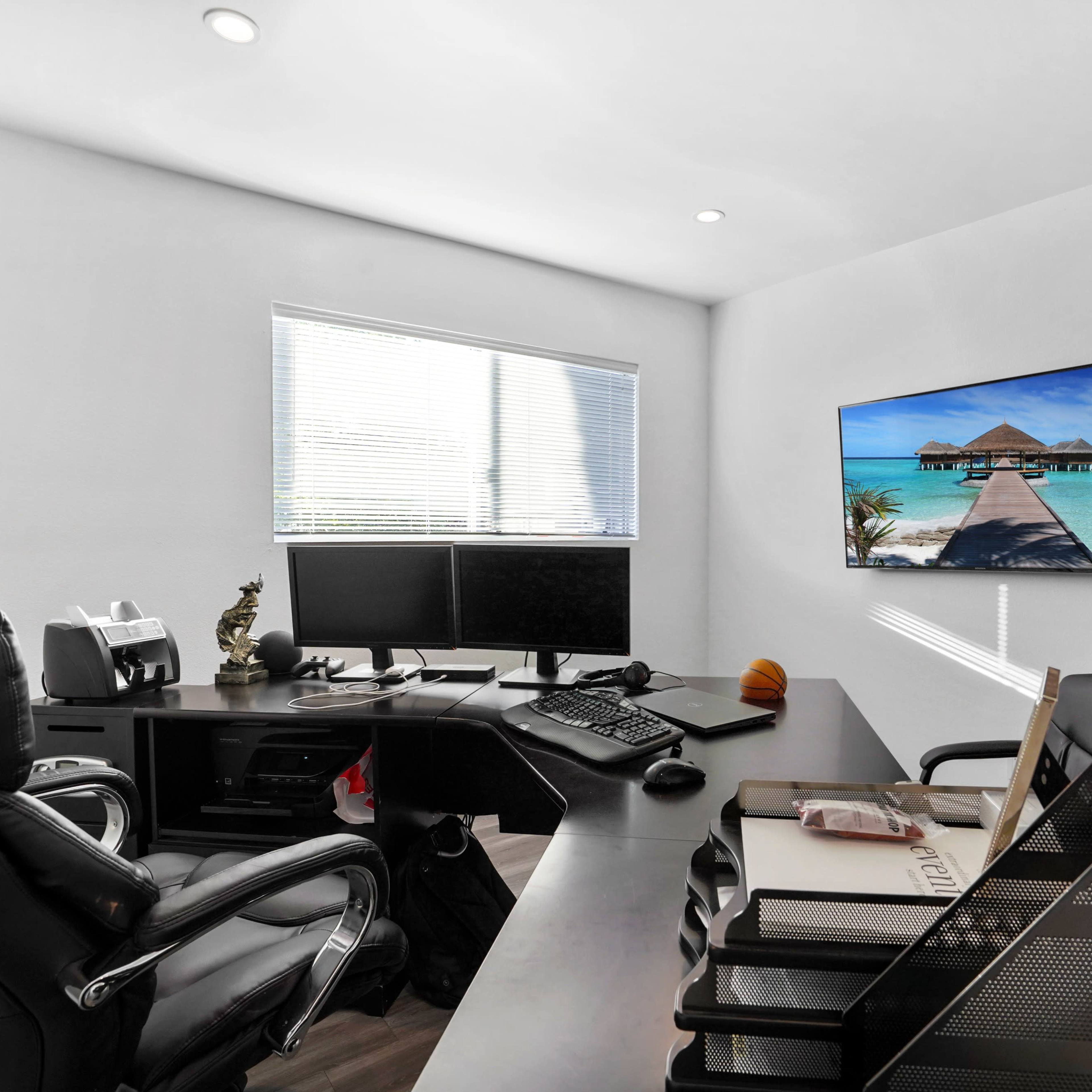 A modern office space with a black leather chair, two computer monitors on a desk, and a television mounted on the wall displaying a beach scene.
