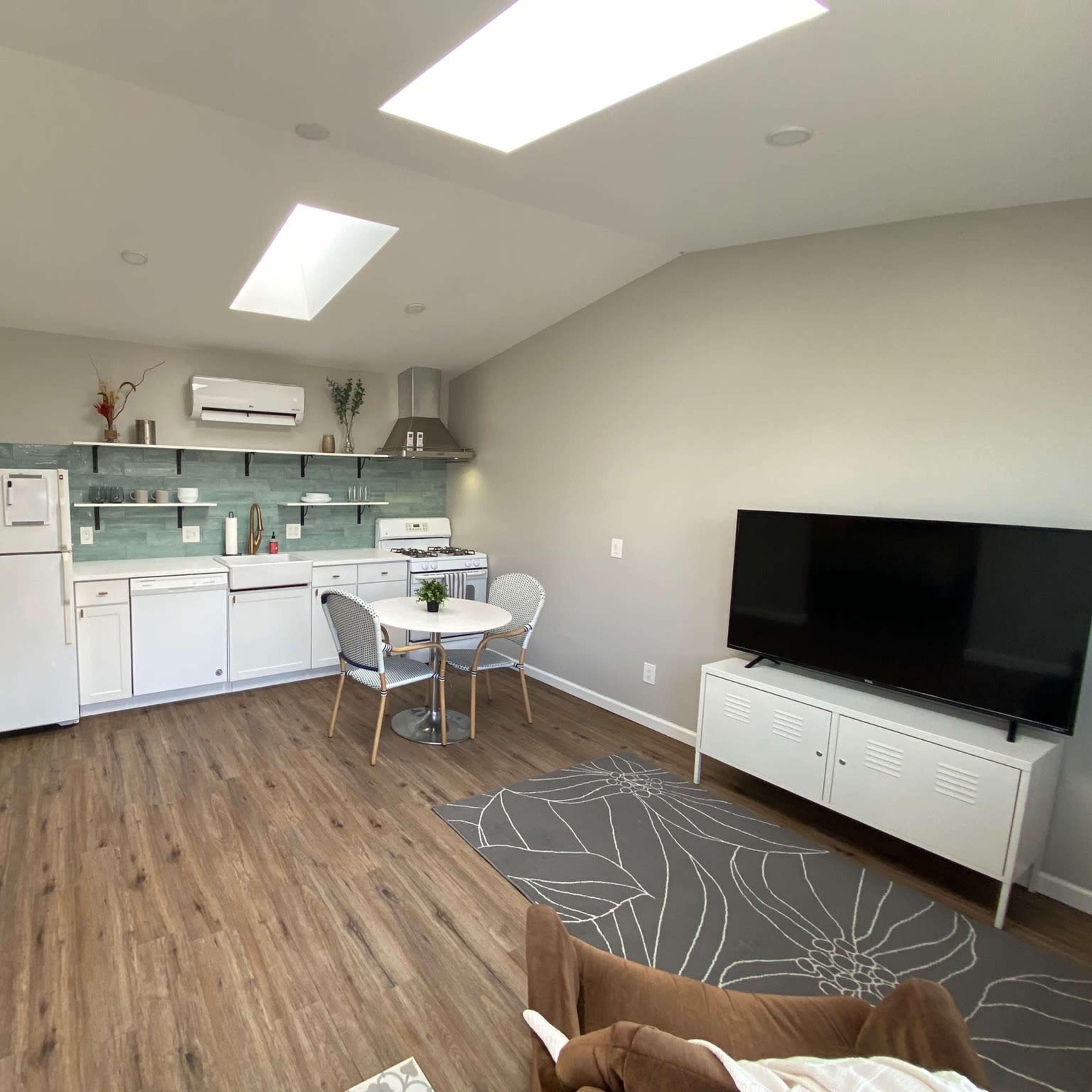 A modern kitchen and living area is shown, featuring a dining table with chairs, a refrigerator, and a television.
