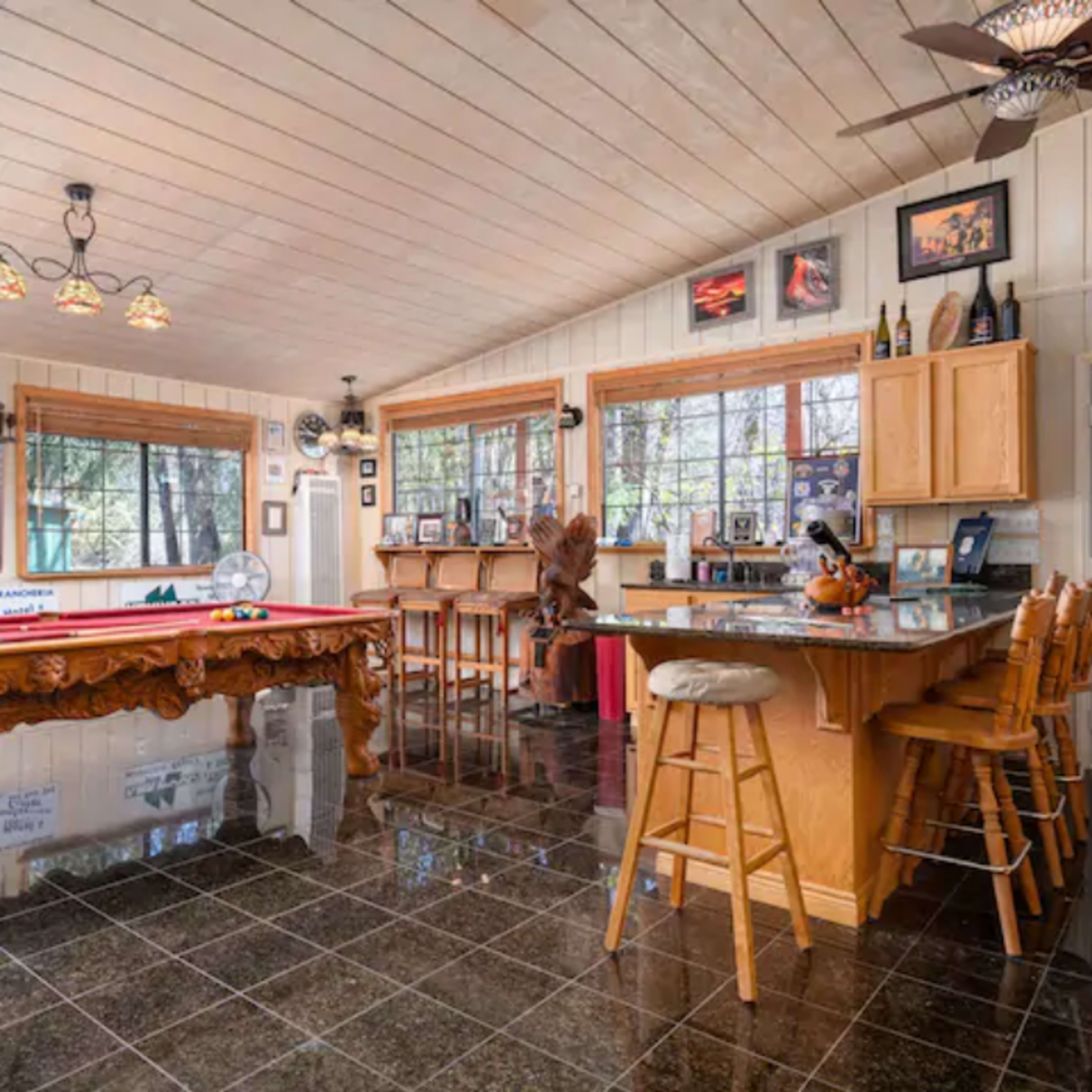 The room features a bar area with wooden stools, a pool table, and large windows that allow natural light to fill the space.