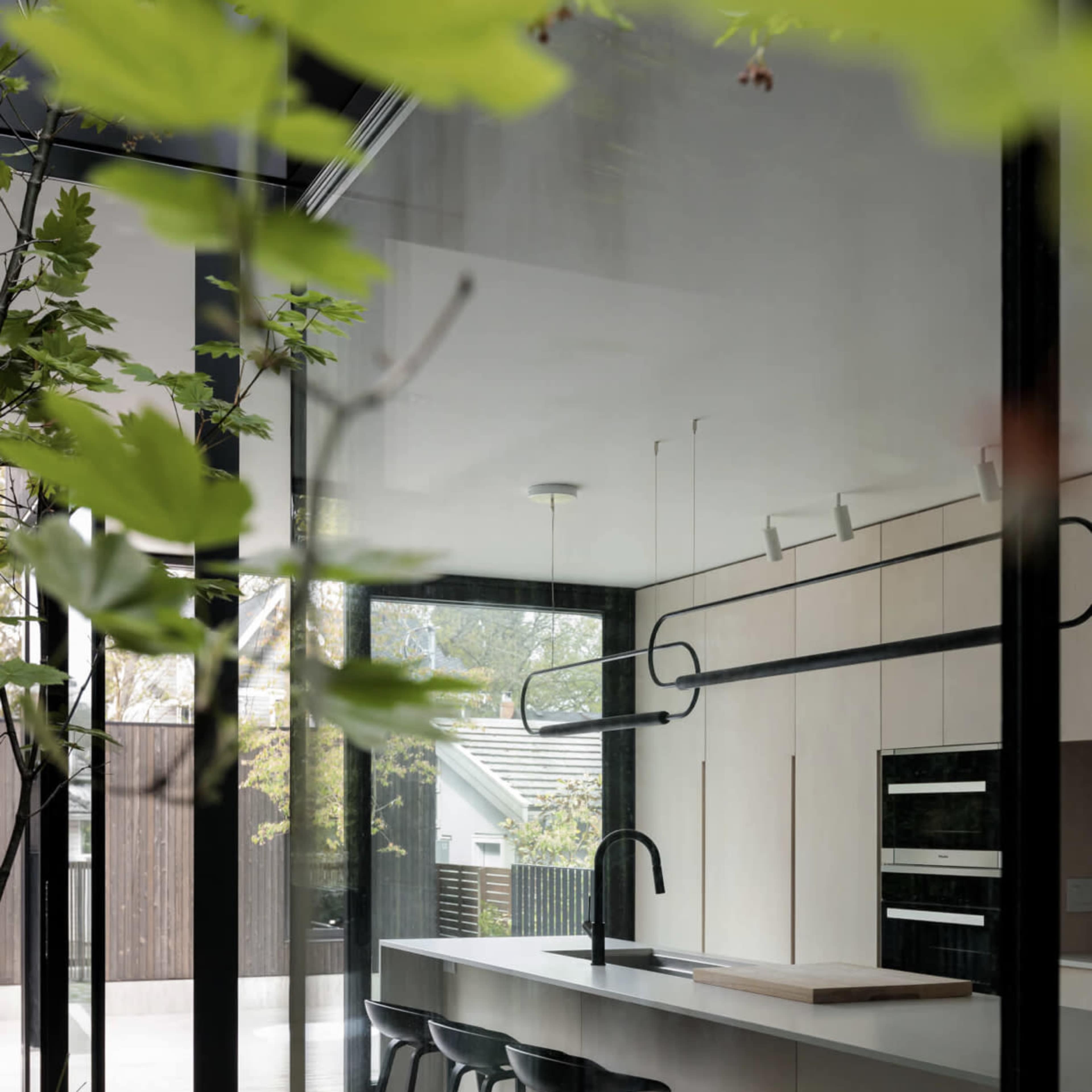 A modern kitchen with a minimalist design featuring a countertop, stools, and large glass windows surrounded by greenery.
