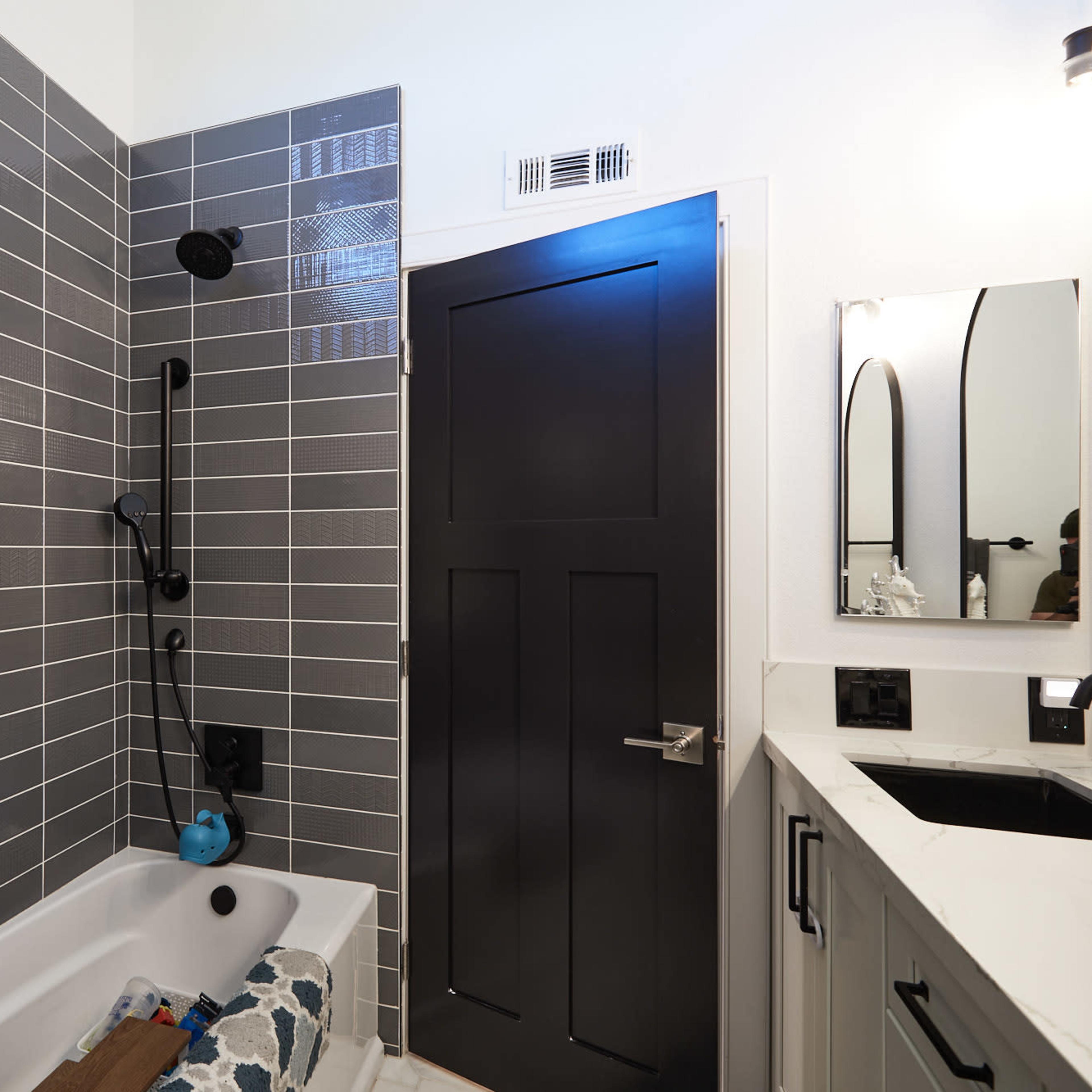 A modern bathroom featuring gray tiled walls, a bathtub with a shower fixture, a dark door, and a countertop with a mirror.