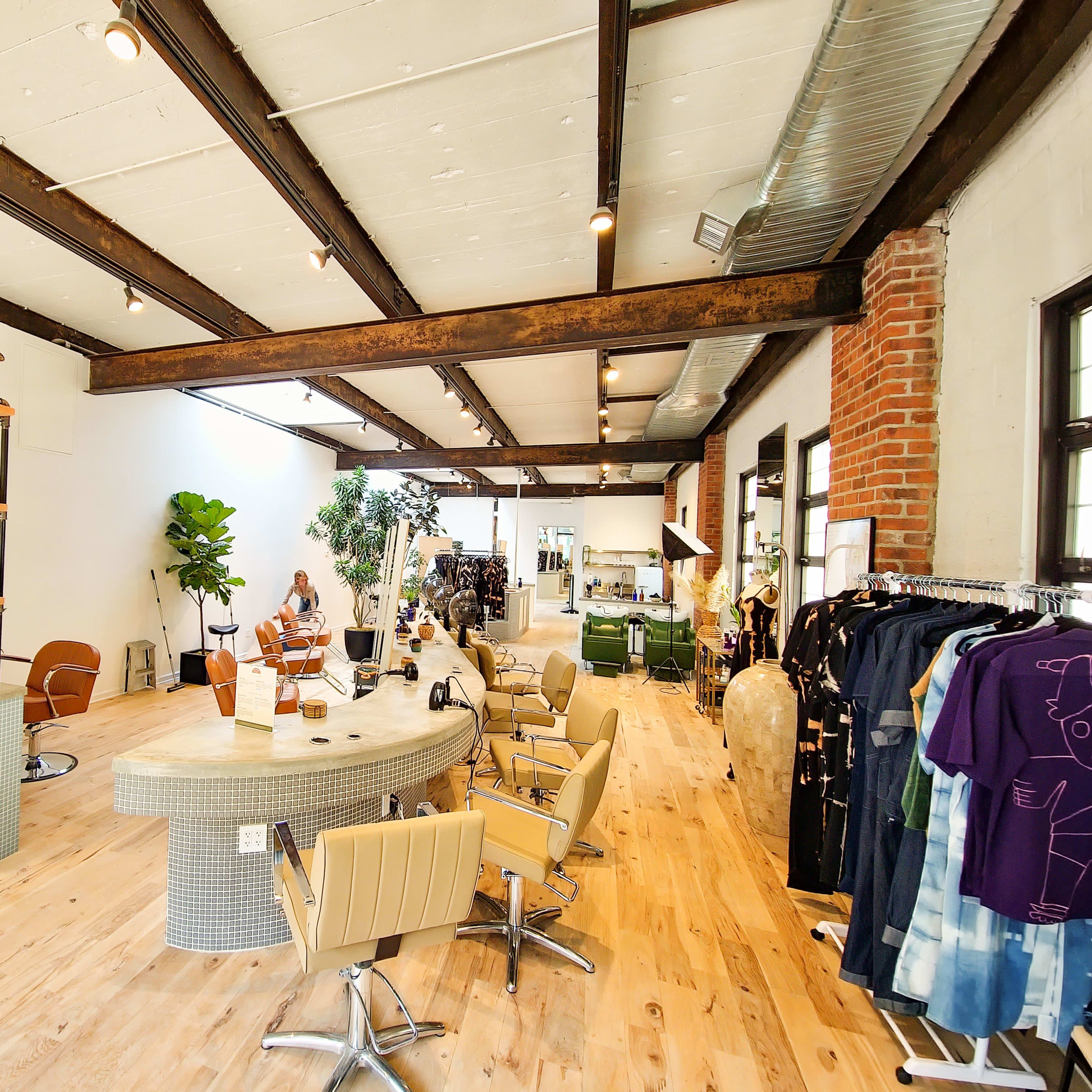 A modern salon interior with a circular reception desk, several styling chairs, plants, and various products displayed on shelves.