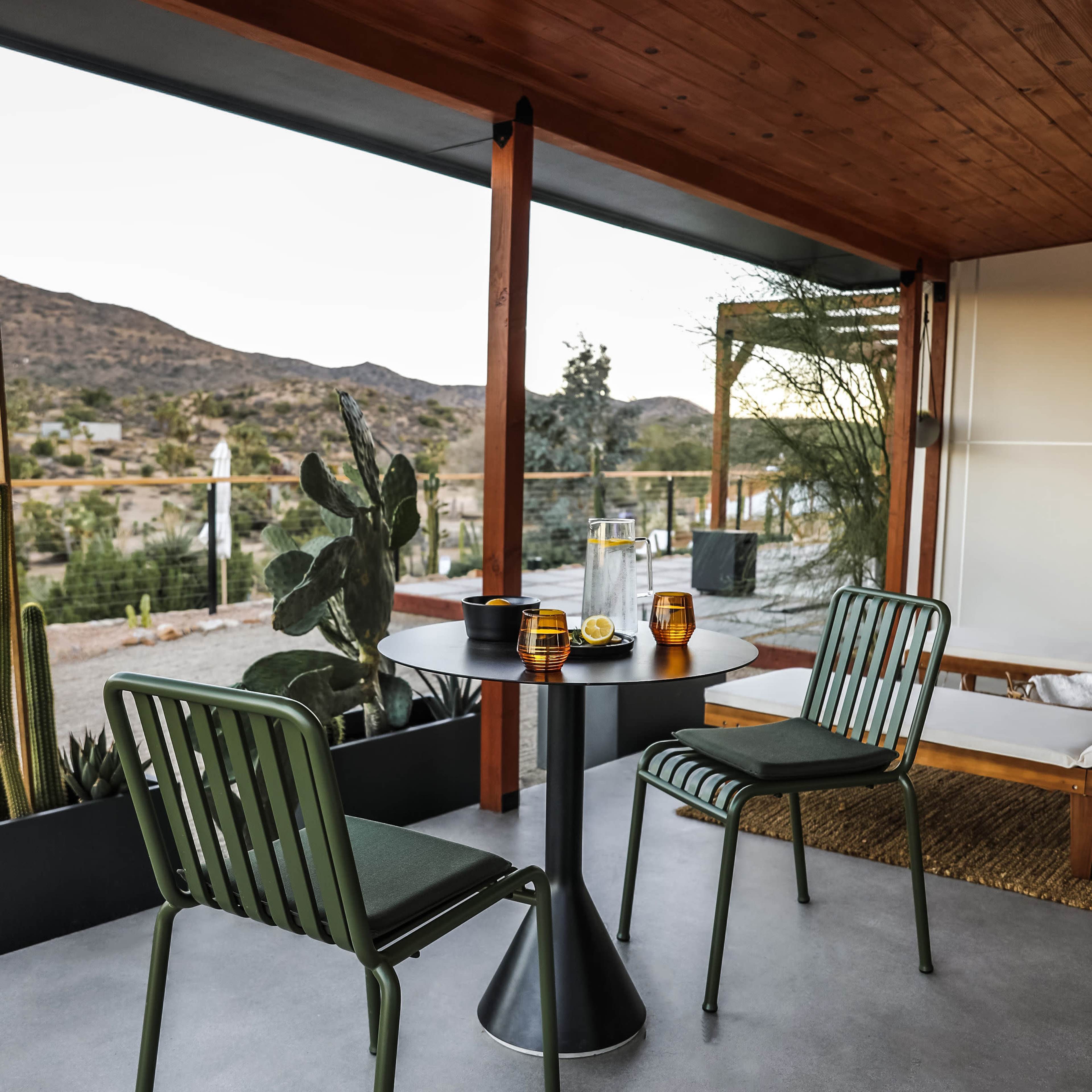 A modern outdoor seating area features two green chairs and a circular table, set against a backdrop of mountains and desert vegetation.