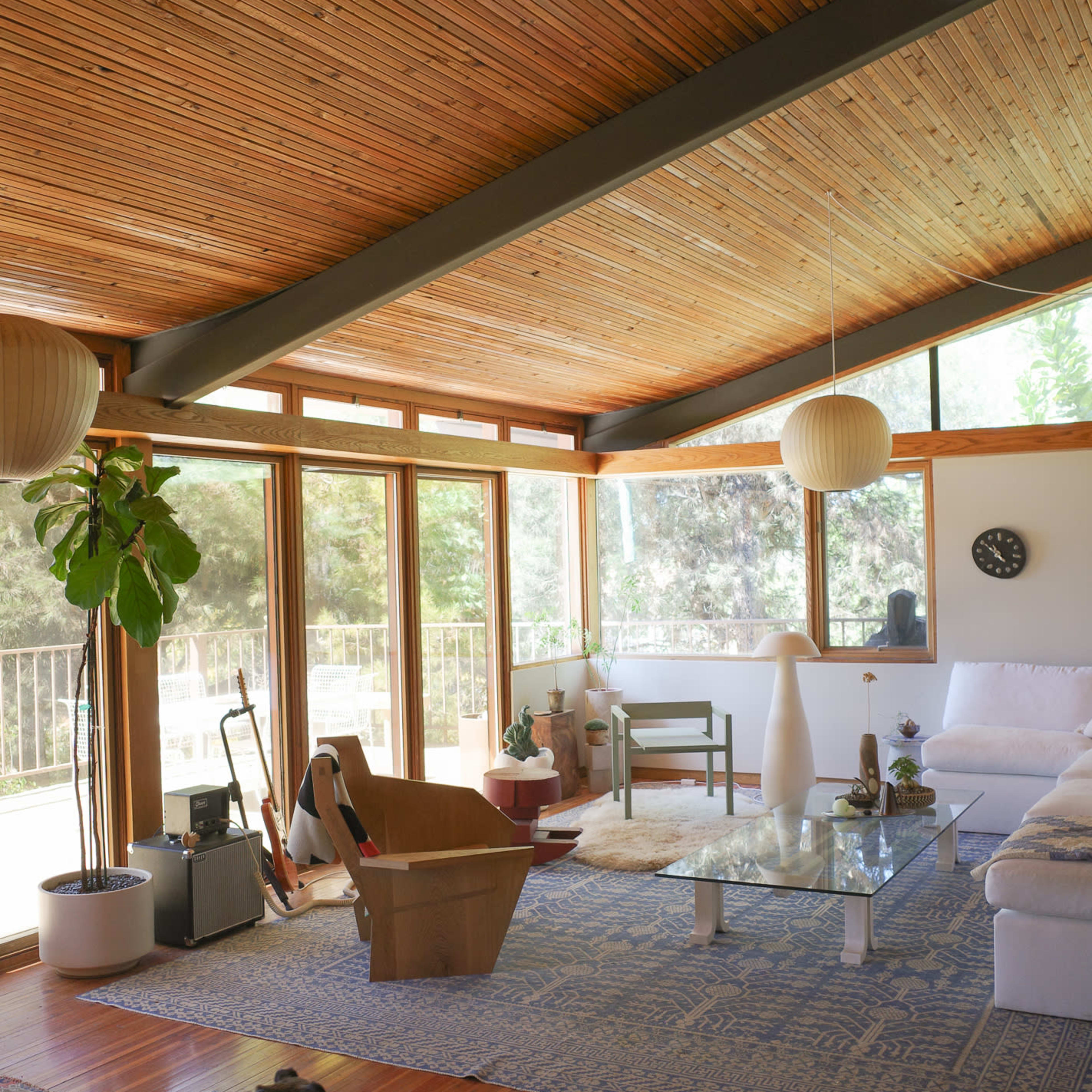 A modern living room with a wooden ceiling, large windows, minimalist furniture, and a dog walking on a rug.
