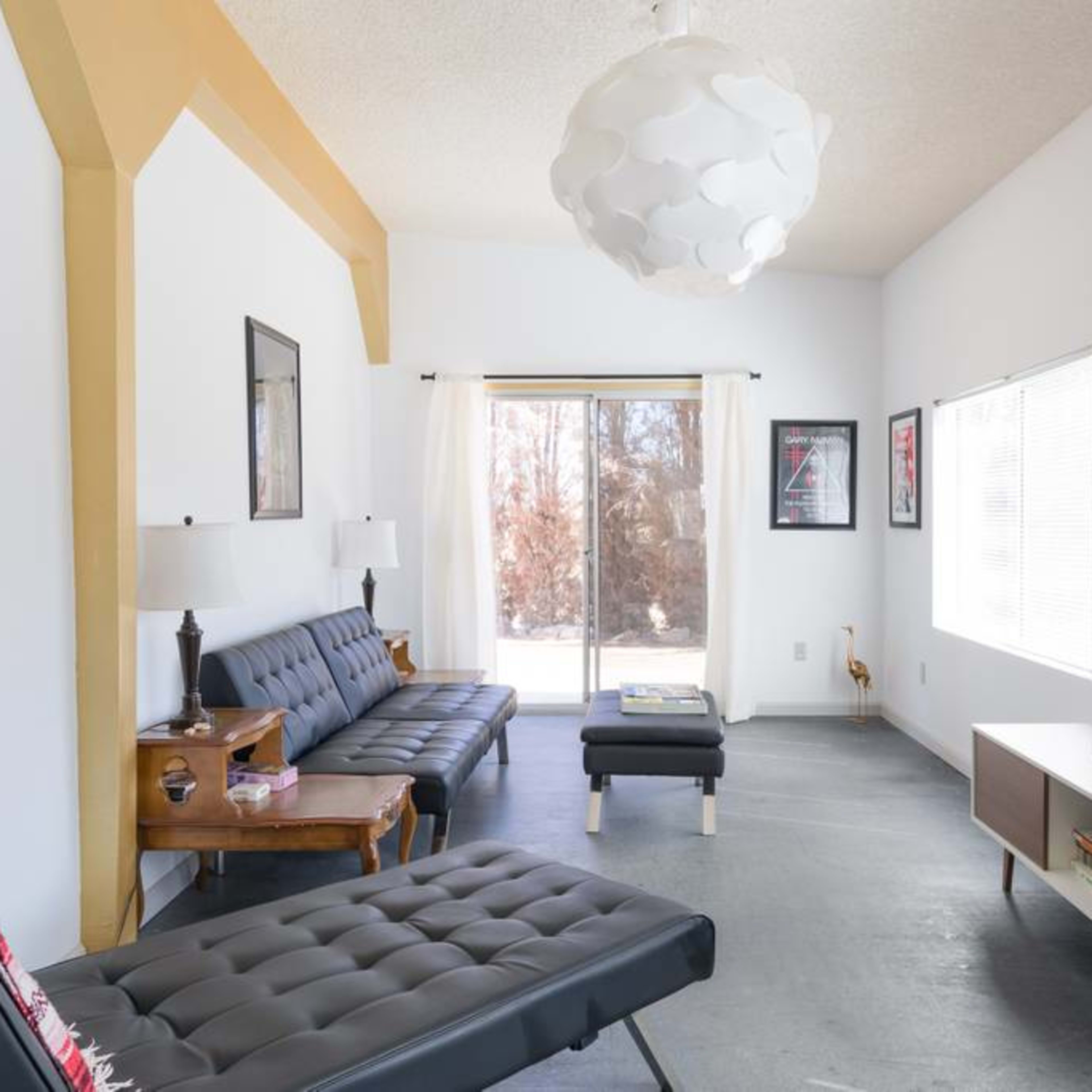 A modern living room features a sleek black sofa, a coffee table, a wall-mounted television, and large windows that provide natural light.