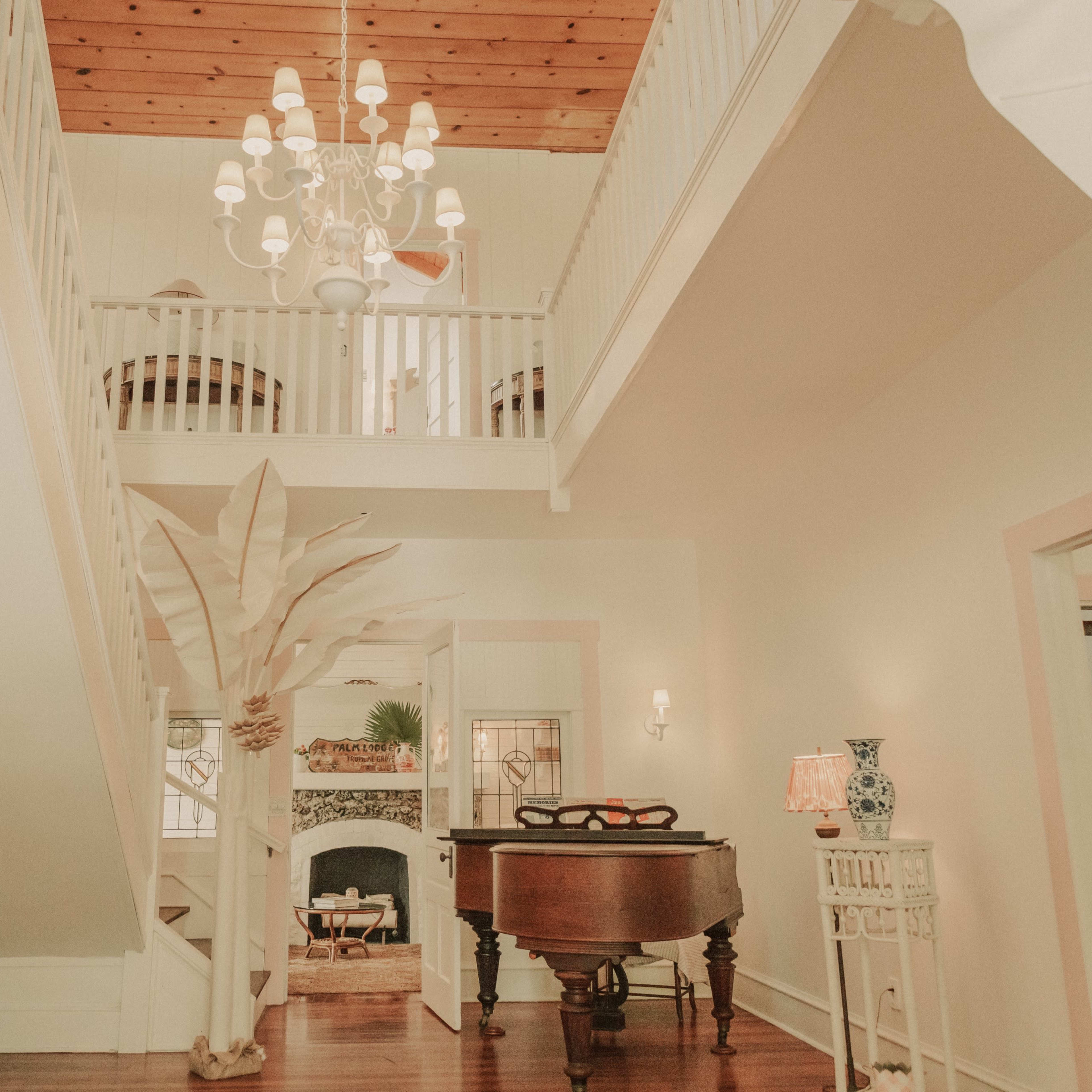 A light-filled entryway with a staircase, a chandelier, a grand piano, and decorative elements like a large plant and a lamp.