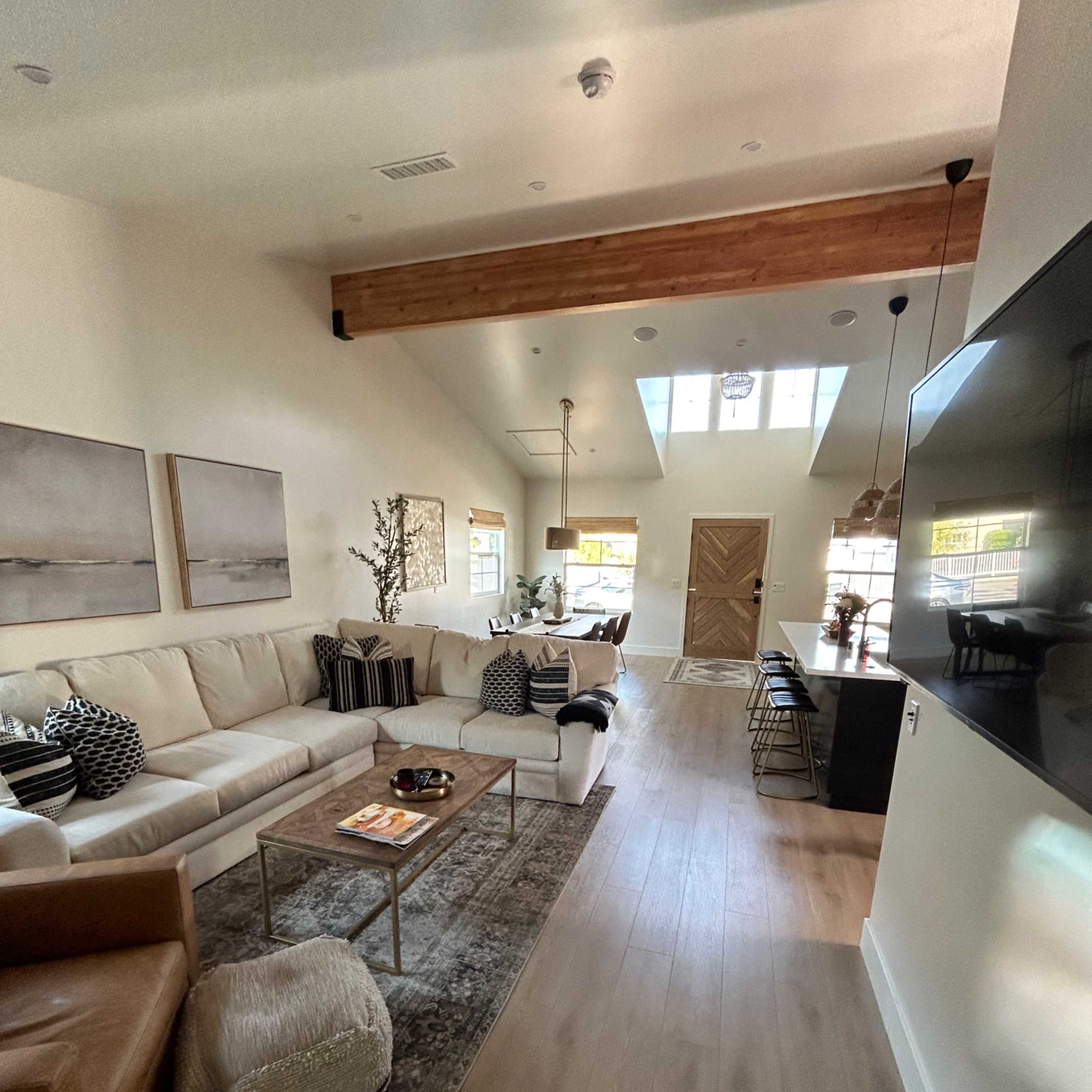 A modern living room with light-colored walls, a large sectional sofa, a coffee table, and a dining area in the background, featuring a high ceiling with wooden beams.