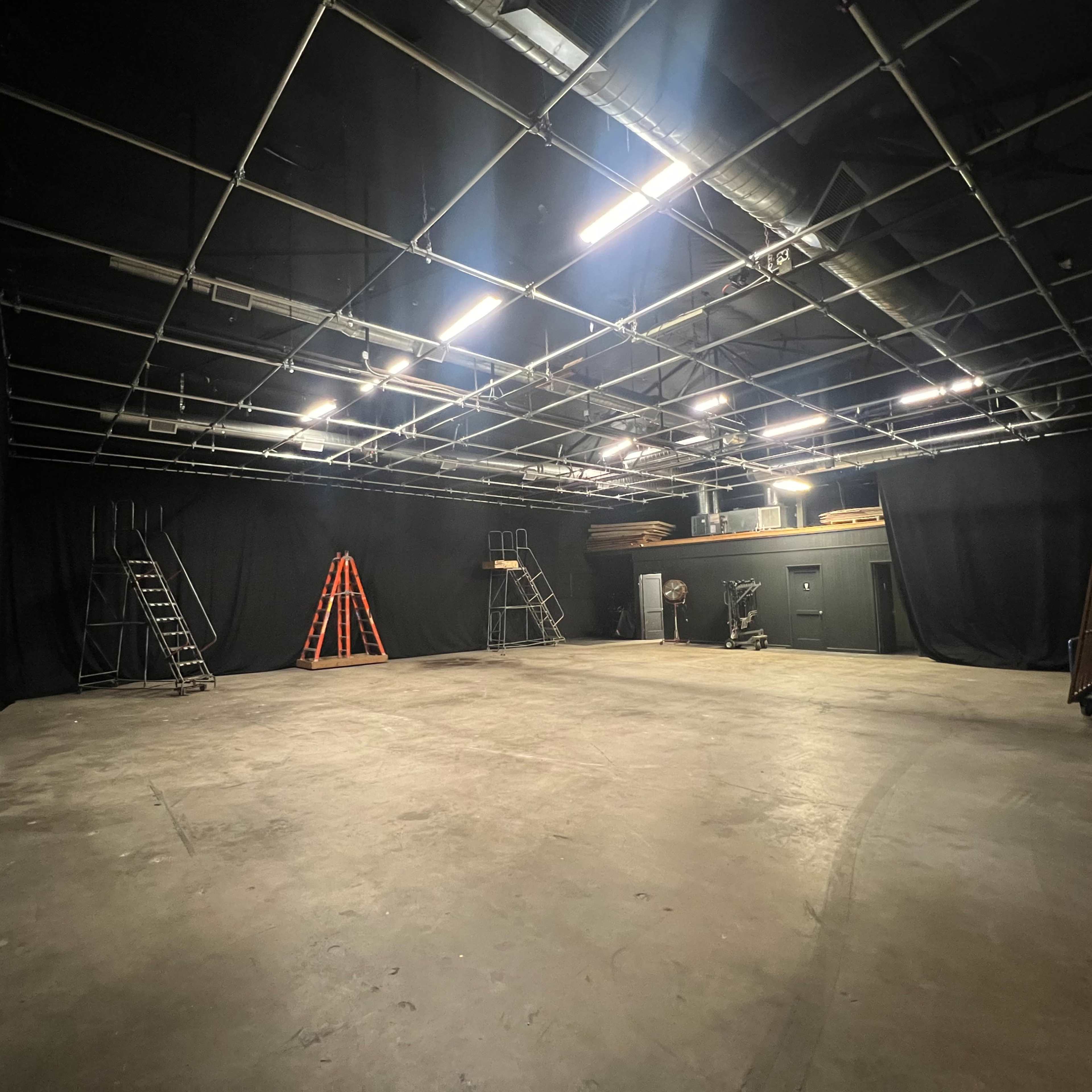 An empty studio space features black curtains lining the walls, several step ladders, and an assortment of equipment arranged in the back.