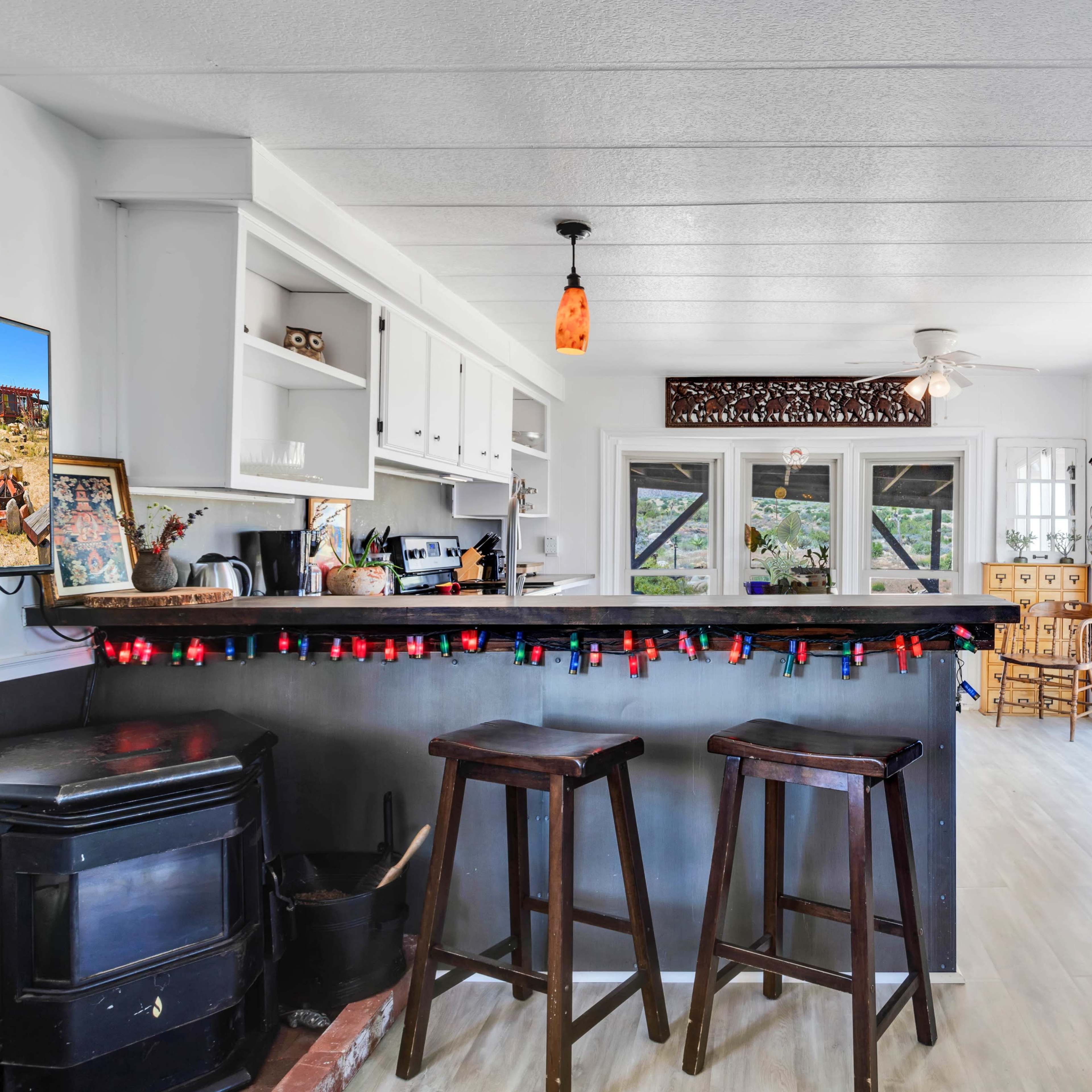 A modern kitchen features a dark countertop with two stools, a television on the wall, and a dining area visible through large windows.