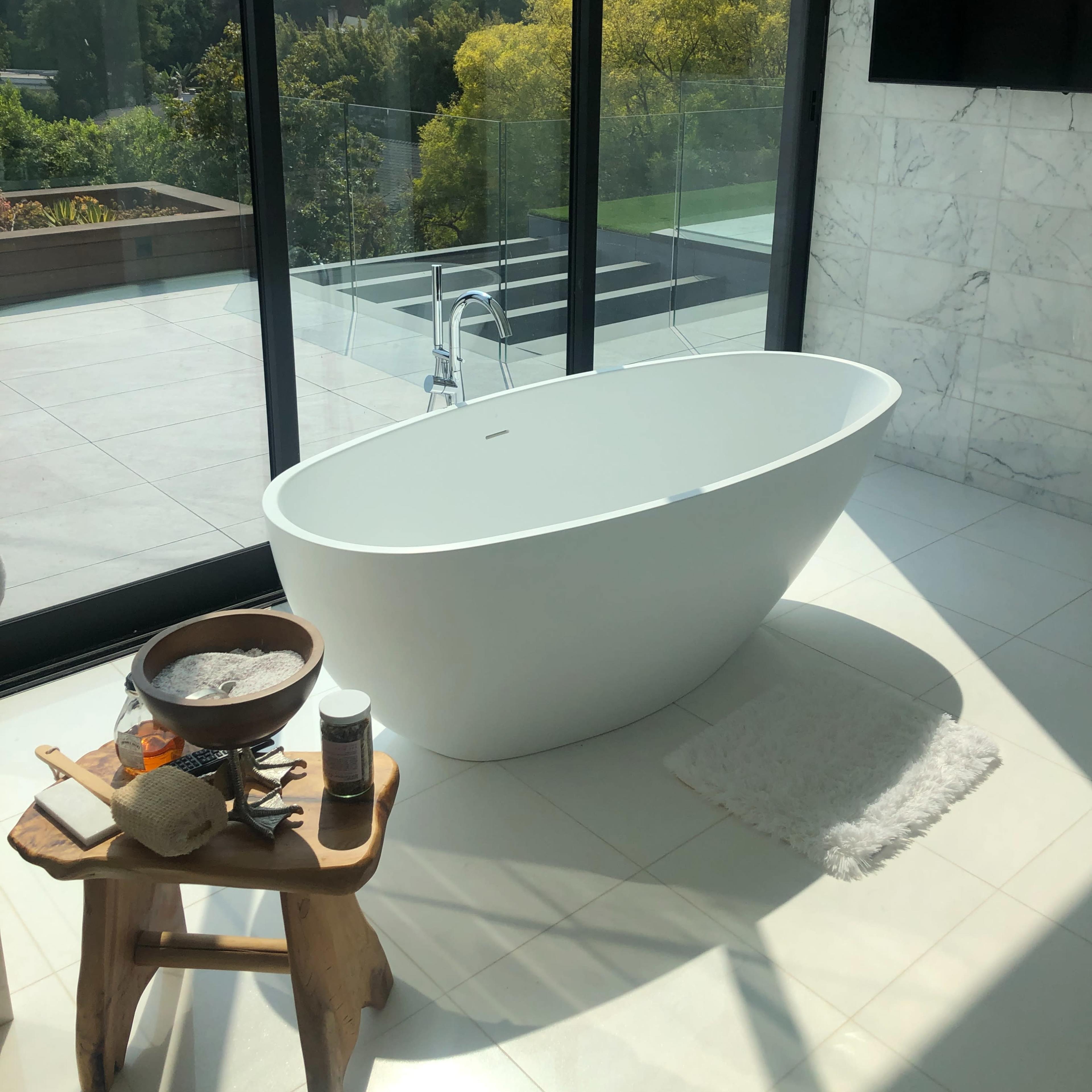 A modern bathroom features a freestanding white bathtub beside a large window that overlooks green vegetation and outdoor landscaping.