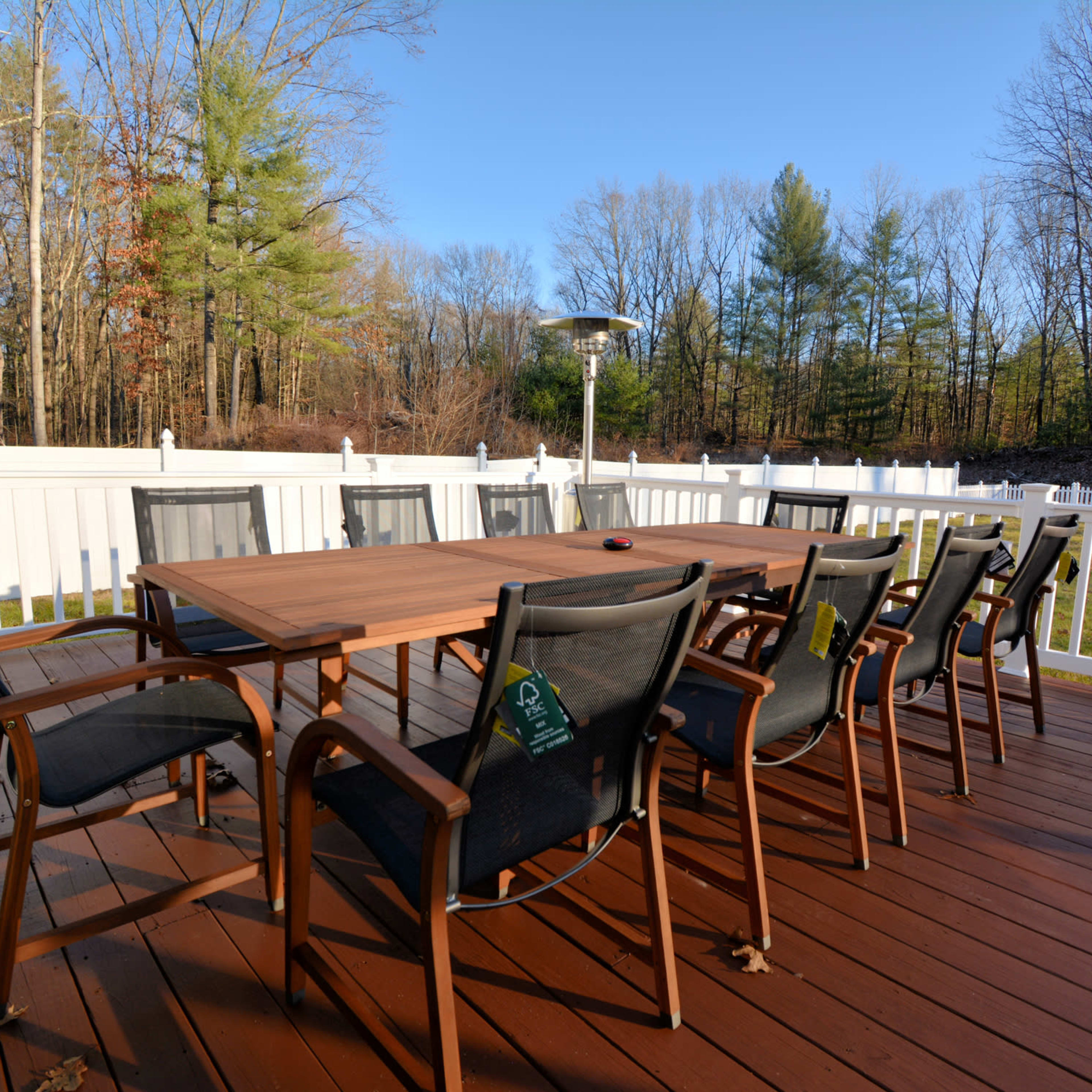 A spacious outdoor patio features a wooden dining table surrounded by several black chairs and a standing heater, set against a backdrop of trees and a white fence.