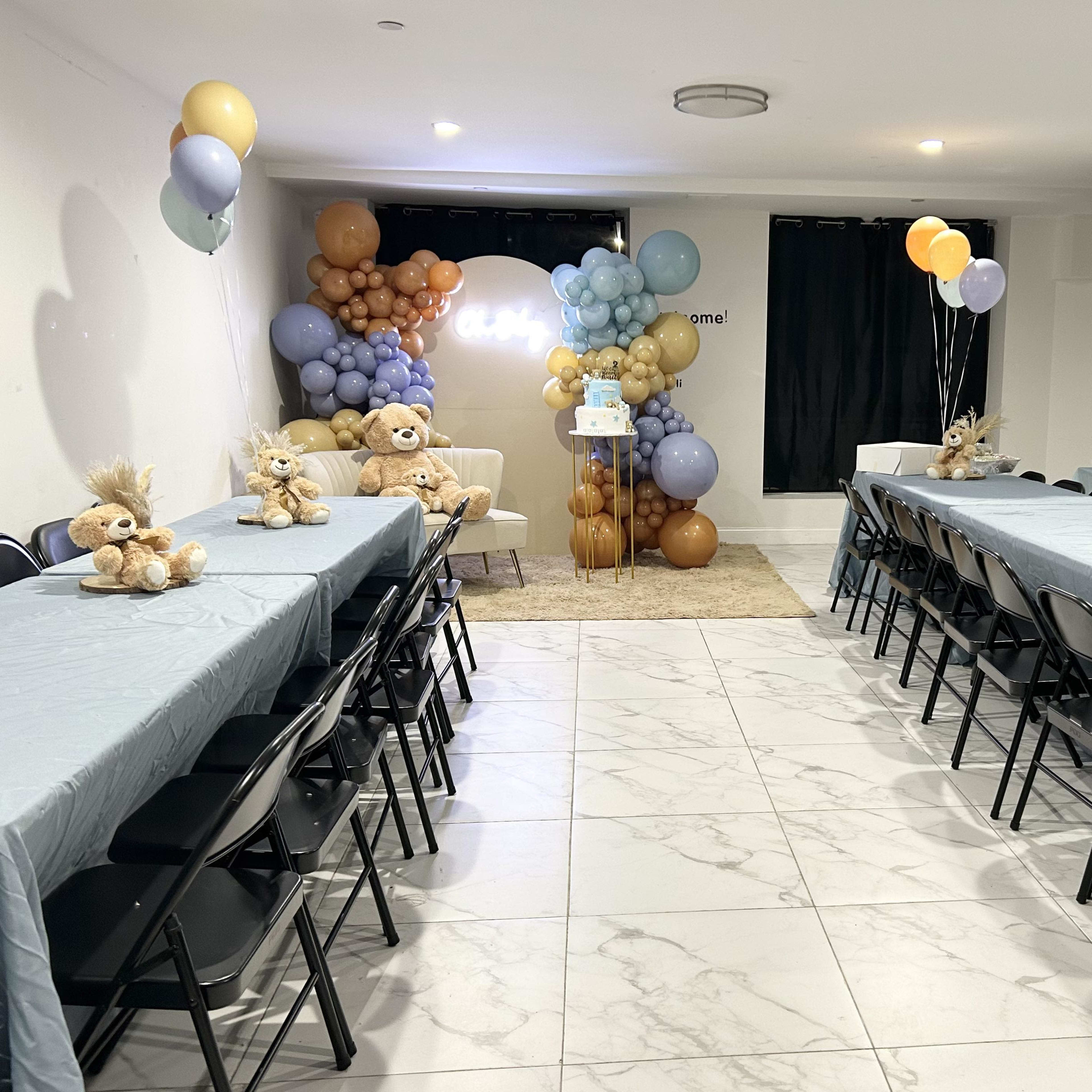 A spacious room is arranged for a party, featuring two long tables covered with light blue tablecloths, teddy bears on each table, and a backdrop of balloon decorations in various colors.