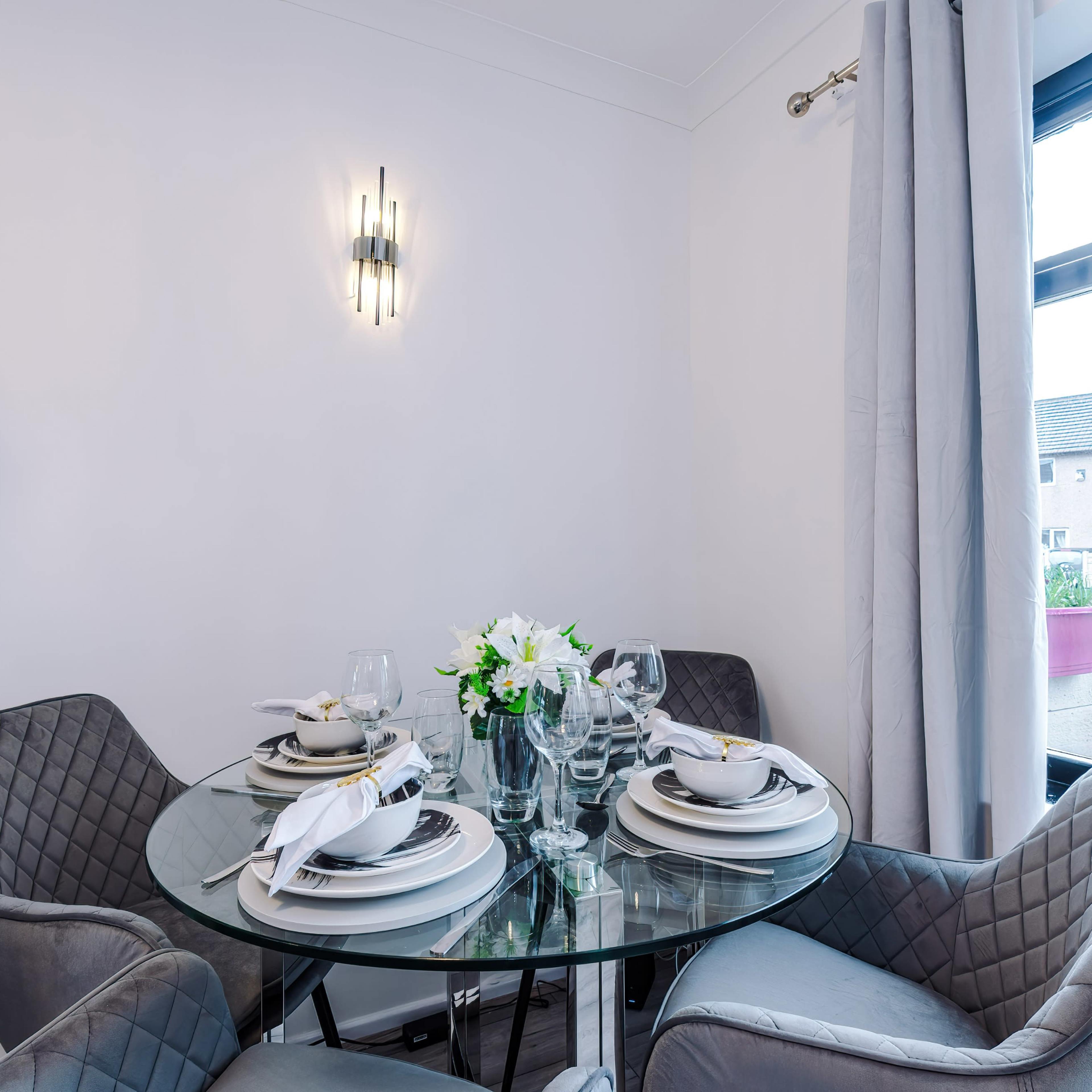 A round glass dining table is set with plates and glasses, surrounded by upholstered chairs in a bright indoor dining area.