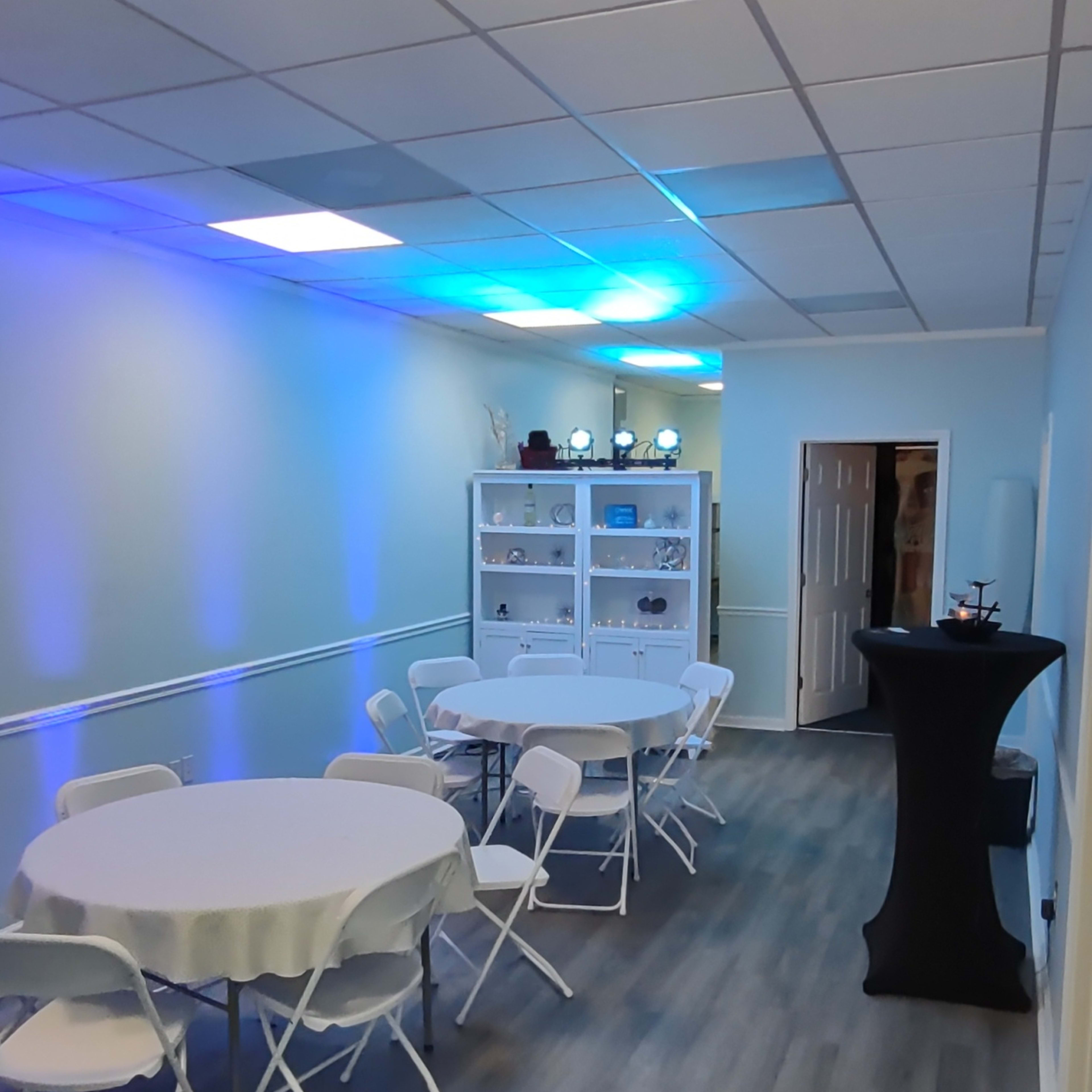 A narrow room with white walls, featuring a round table with chairs, a black high table, and shelves displaying various items, all illuminated by blue lighting.