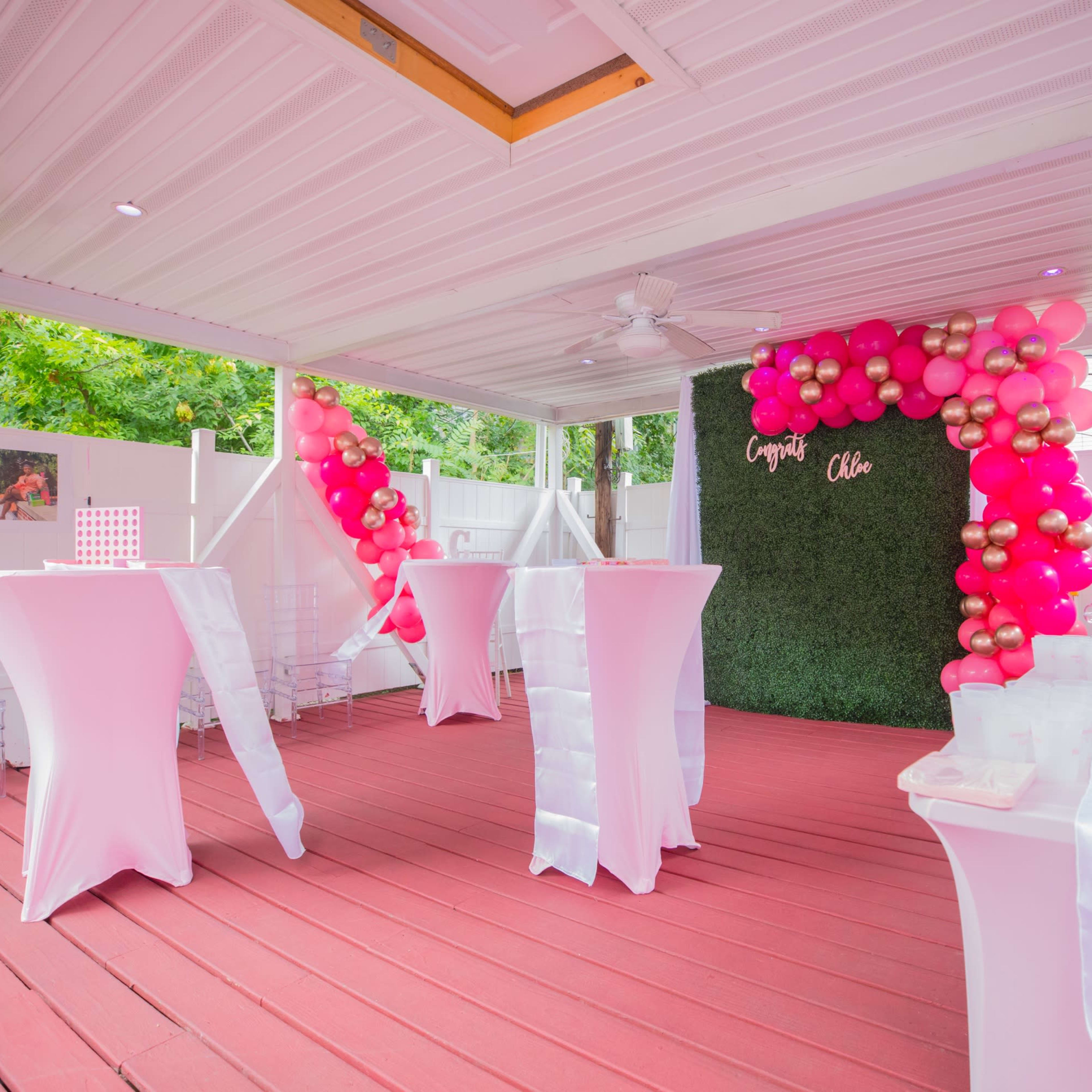 A decorated outdoor event space featuring pink and gold balloon arrangements, white cocktail tables, and a greenery backdrop.