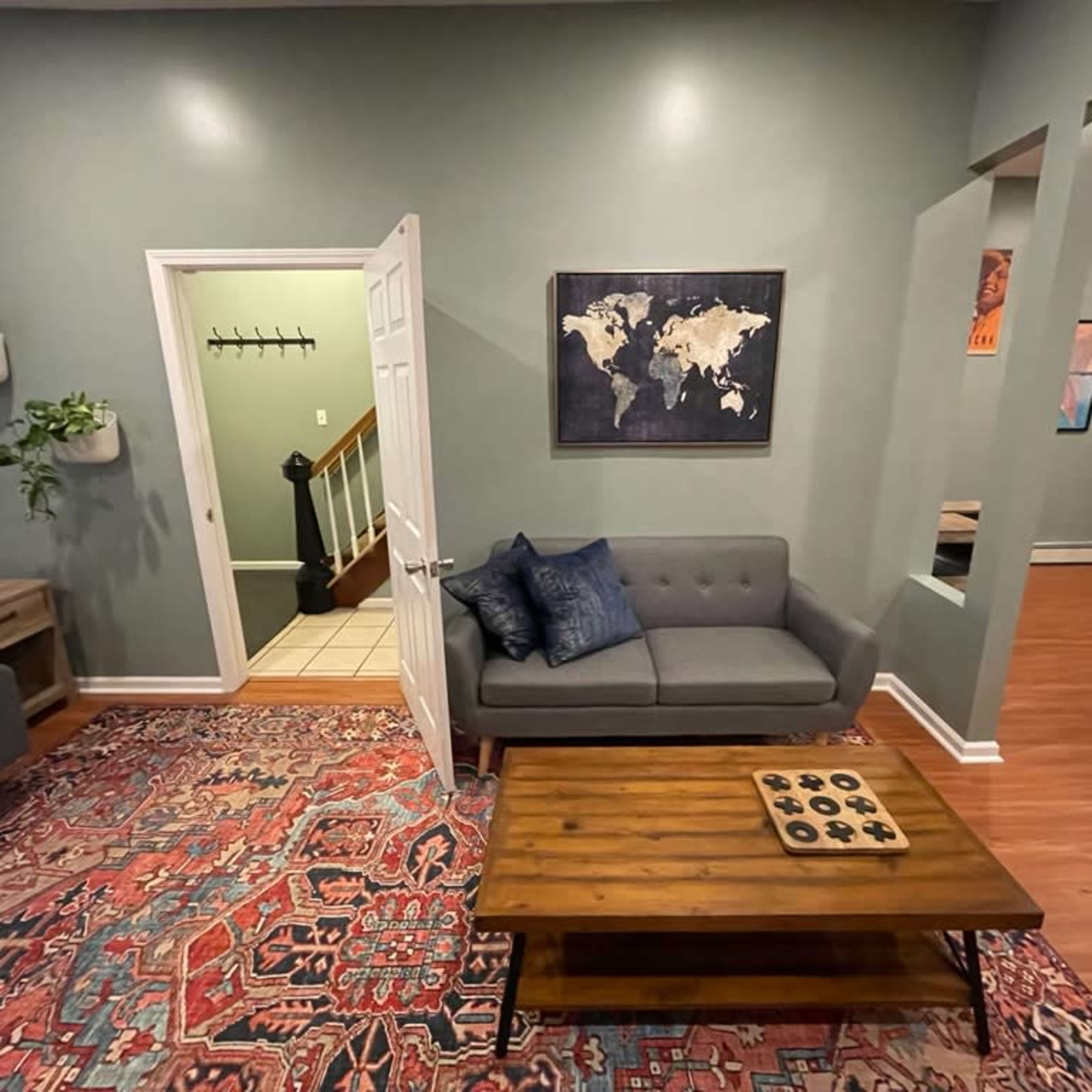 A living room with a gray sofa, a wooden coffee table with a game set, and a map on the wall, leading to a staircase and another room.