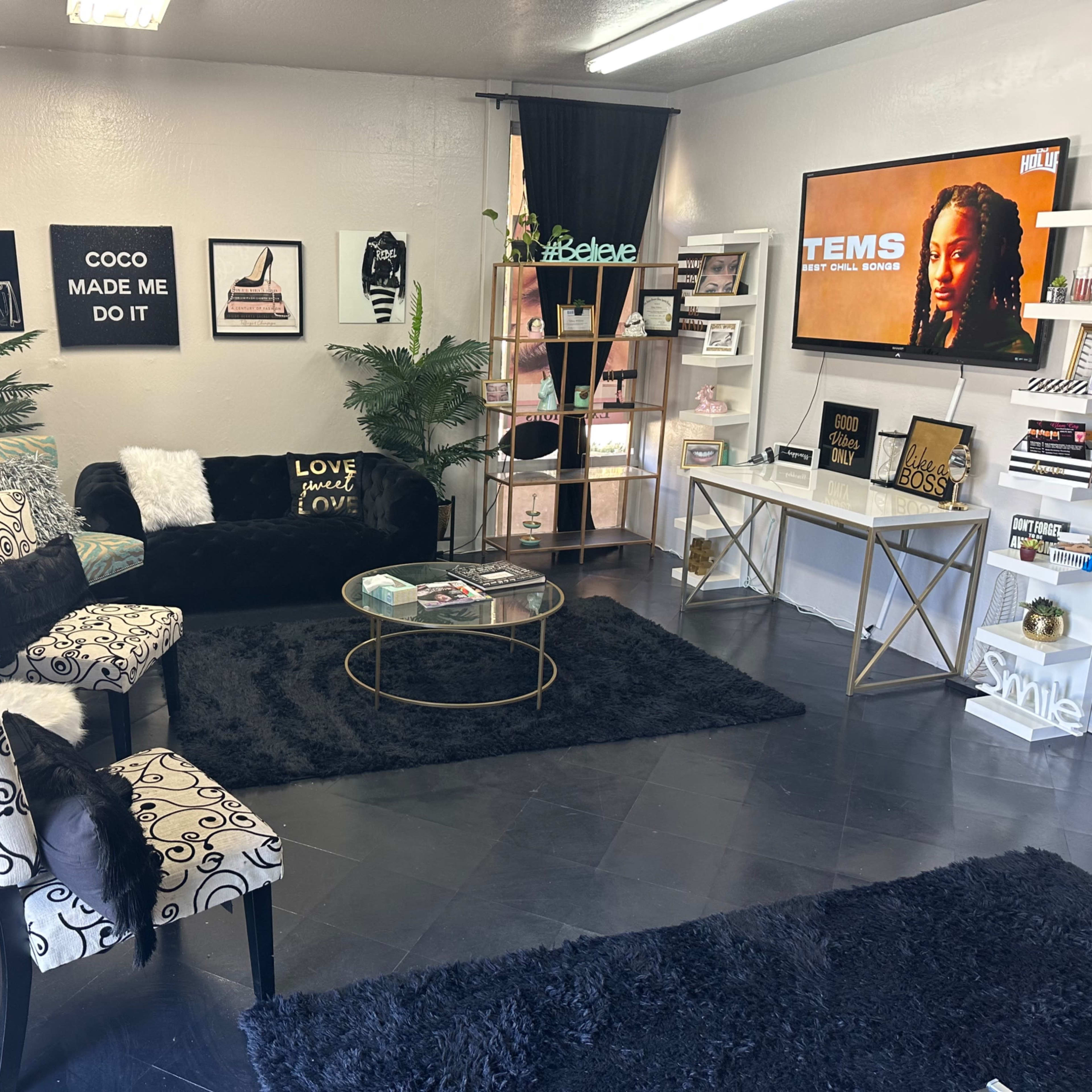 A modern living room featuring a black sofa, patterned chairs, a coffee table, decorative shelves with books and plants, and a television on the wall.