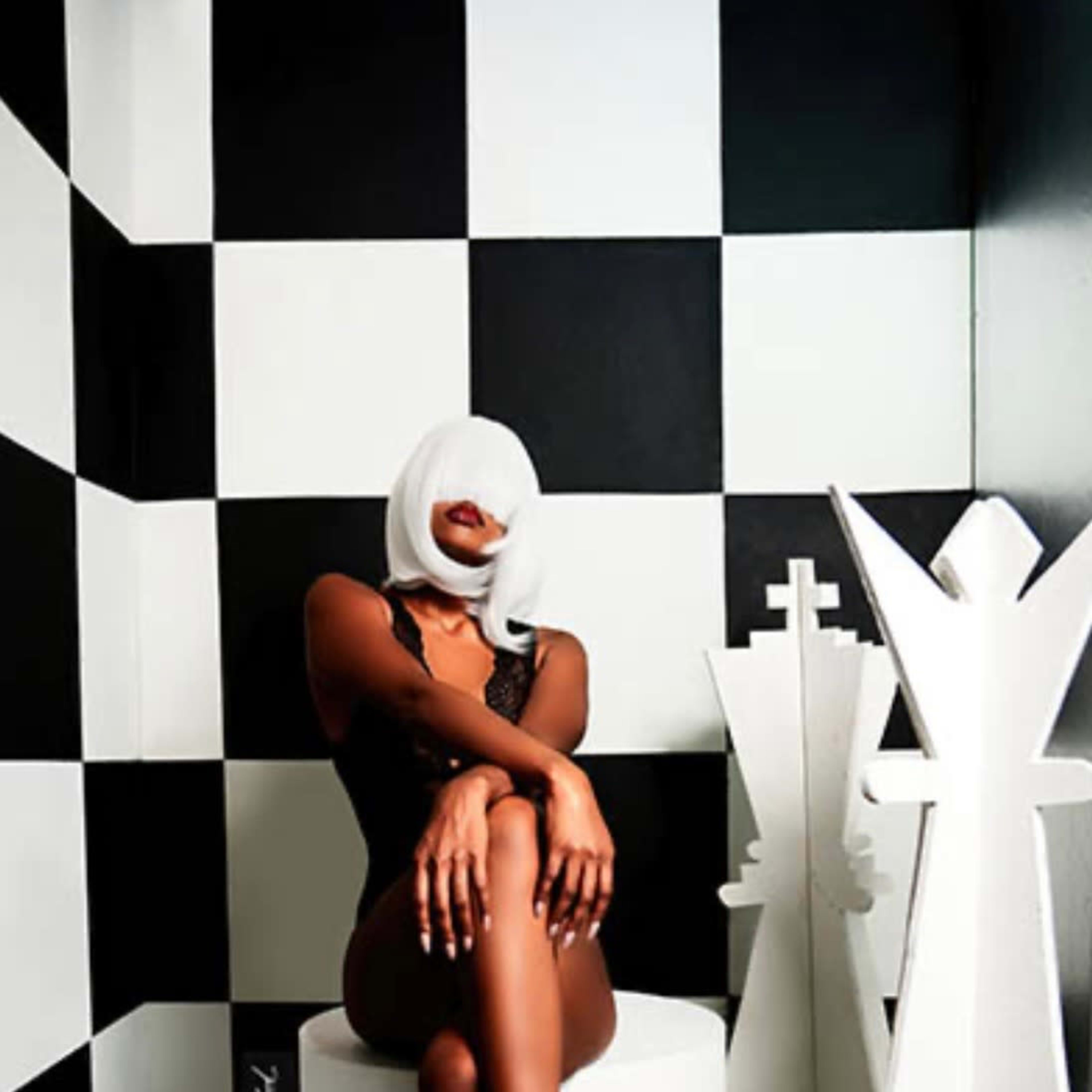 A person with a white headscarf sits elegantly on a white stool in a room with black and white checkered walls, next to a large white chess piece.