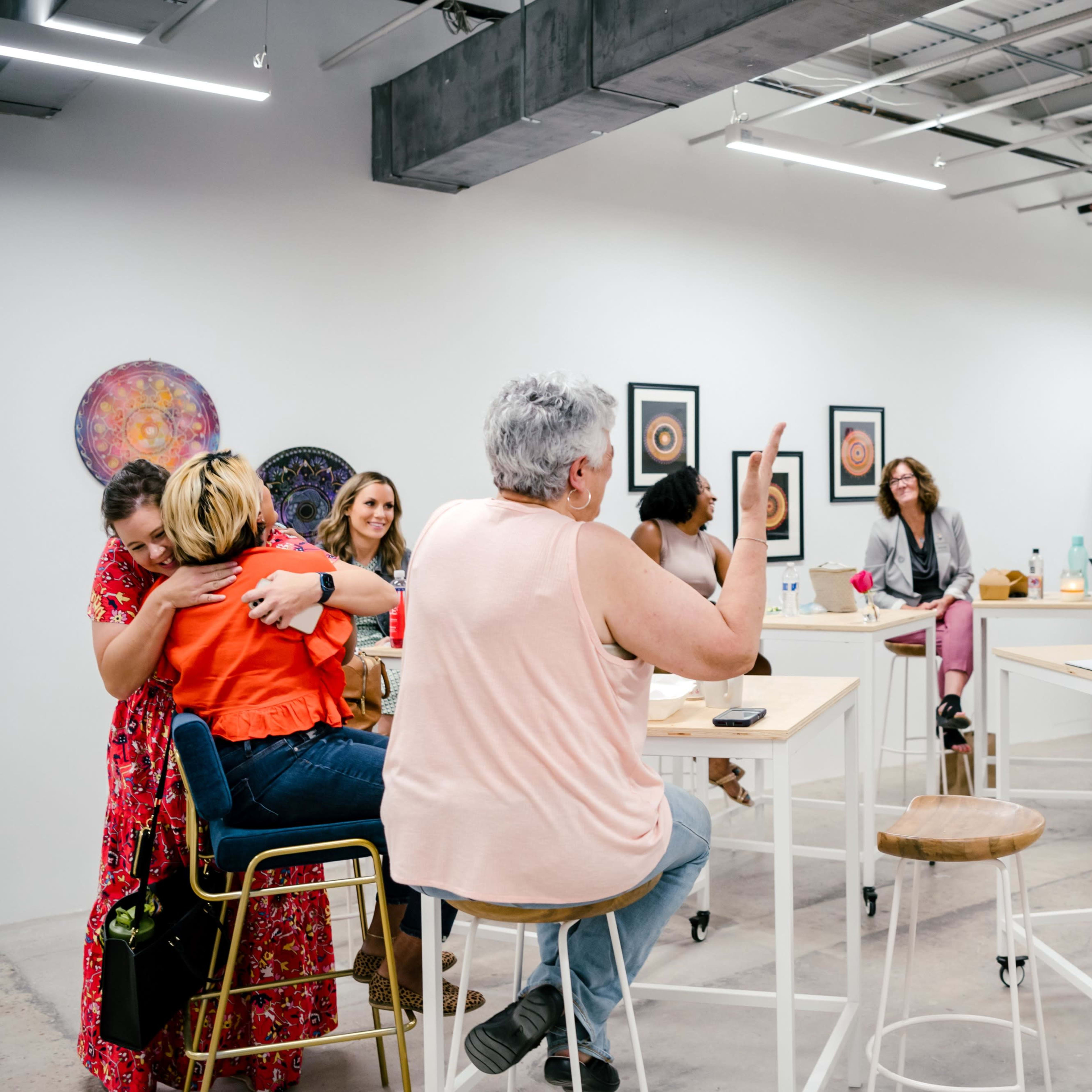 A group of people engages in conversation and laughter in a brightly lit, modern workspace equipped with tall tables and artwork on the walls.