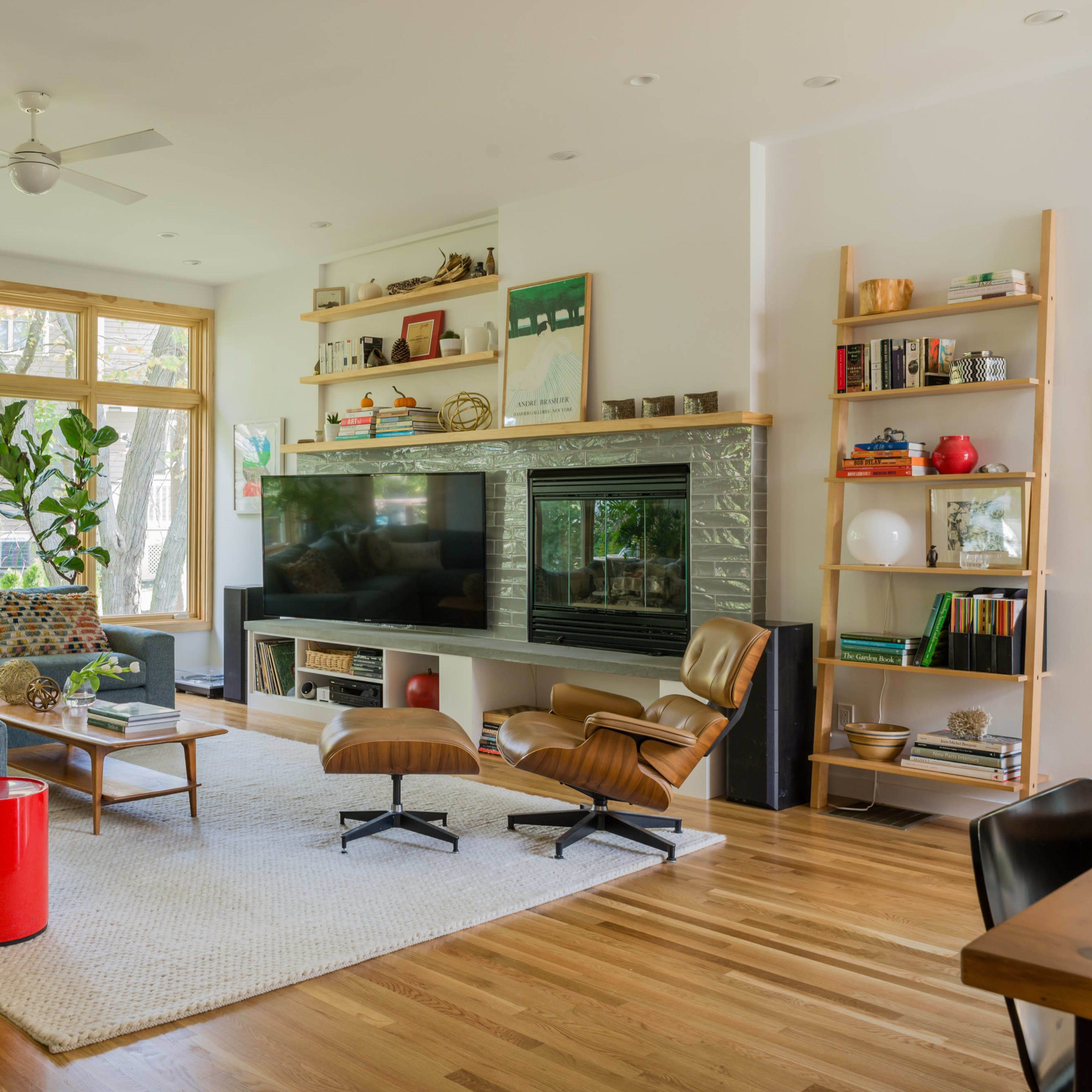 A modern living room features a large sectional sofa, a television set against a stone wall, and a mix of bookshelves and decorative plants near large windows.