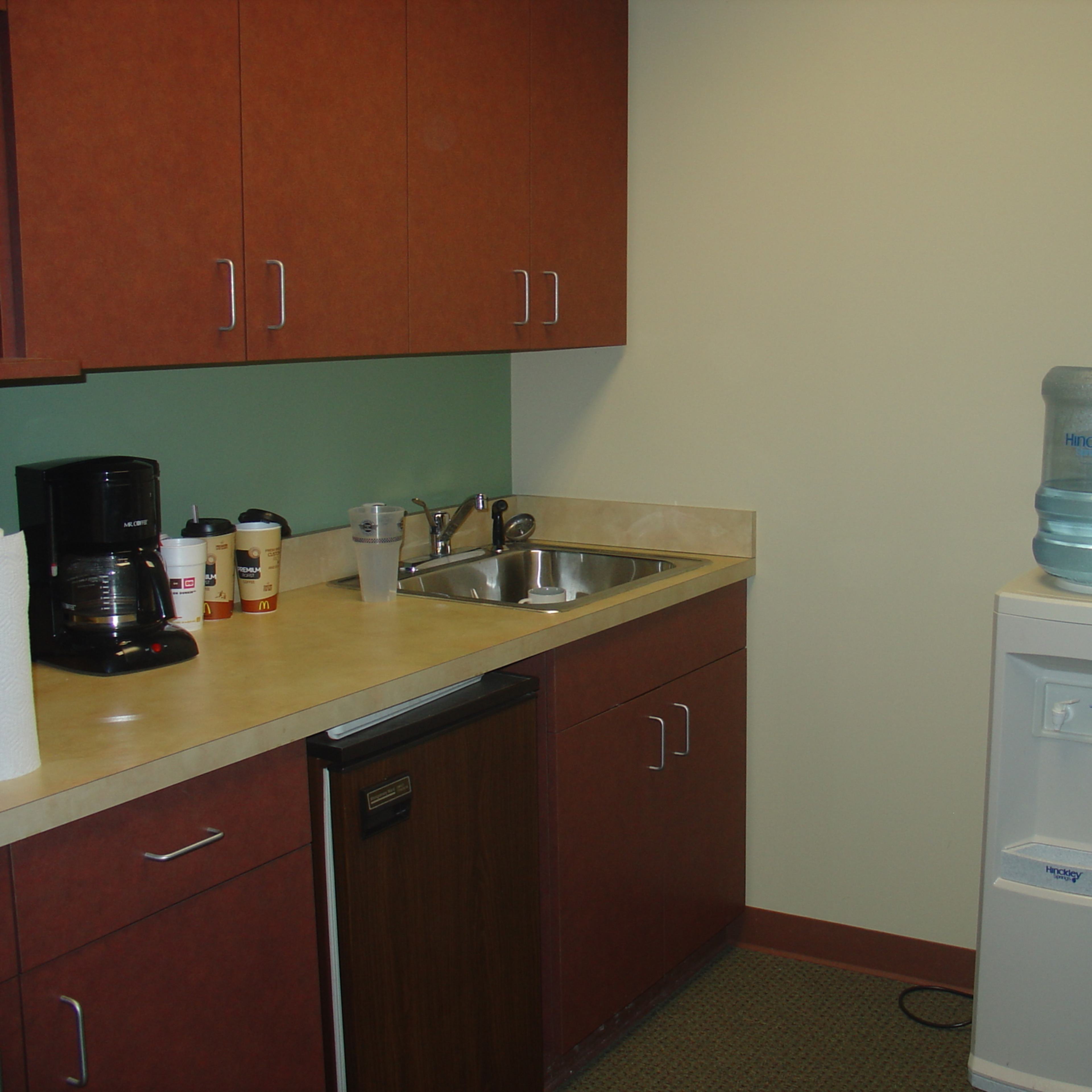 A small kitchen area with wooden cabinets, a sink, a coffee maker, and a water cooler.