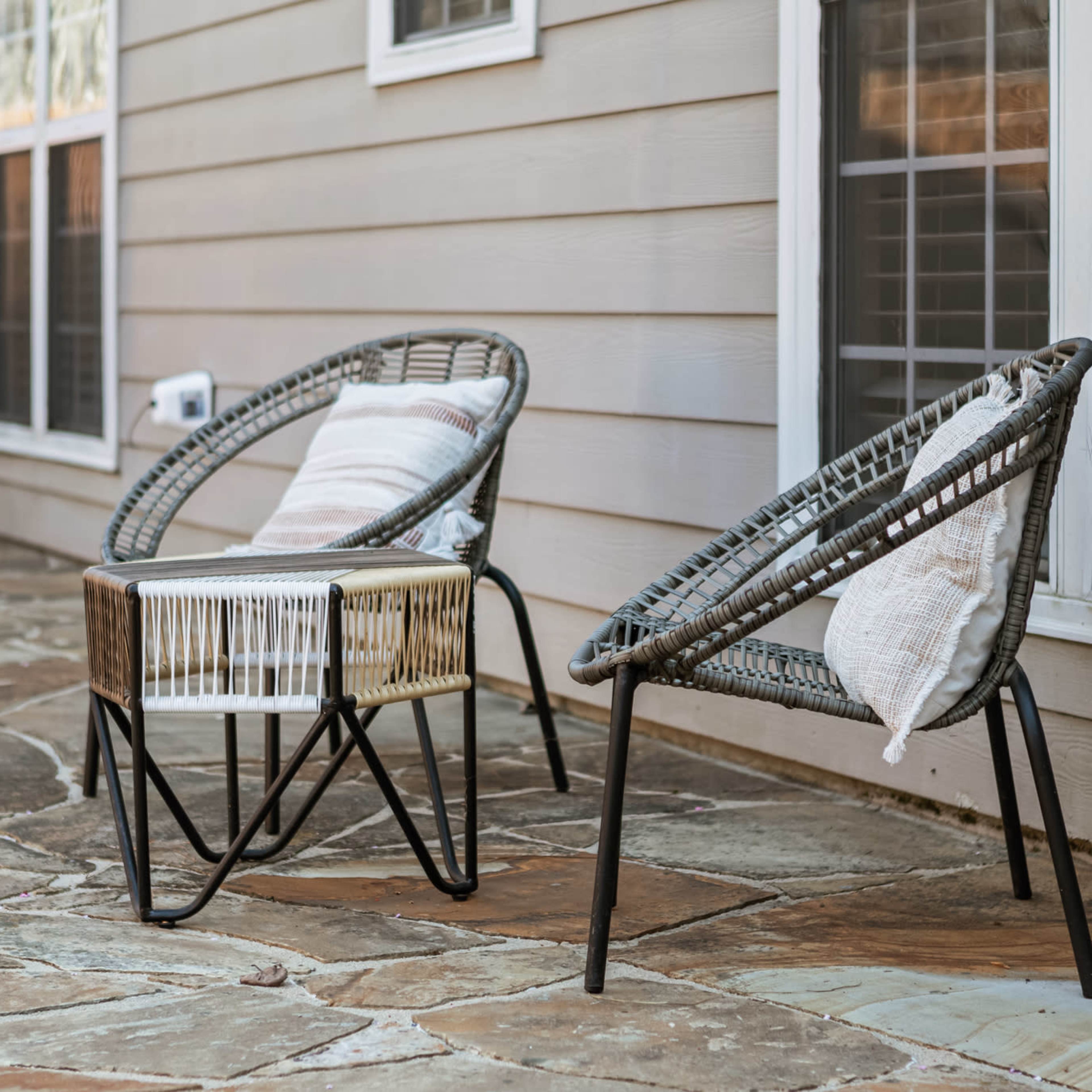 Two wicker chairs and a low table are arranged on a stone patio beside a house with multiple windows.