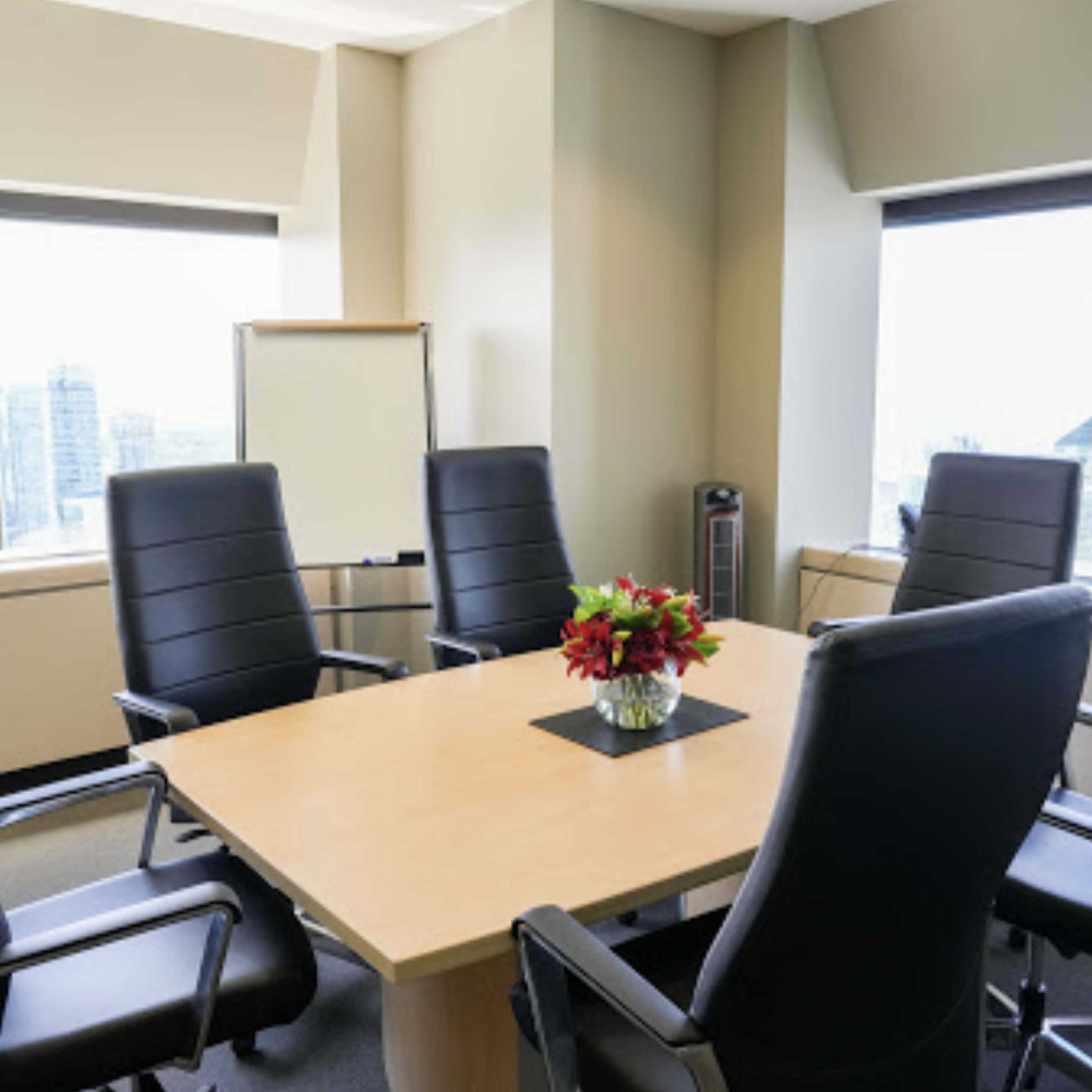 A conference room features a round wooden table surrounded by six black leather chairs, with large windows providing a view of the city skyline.