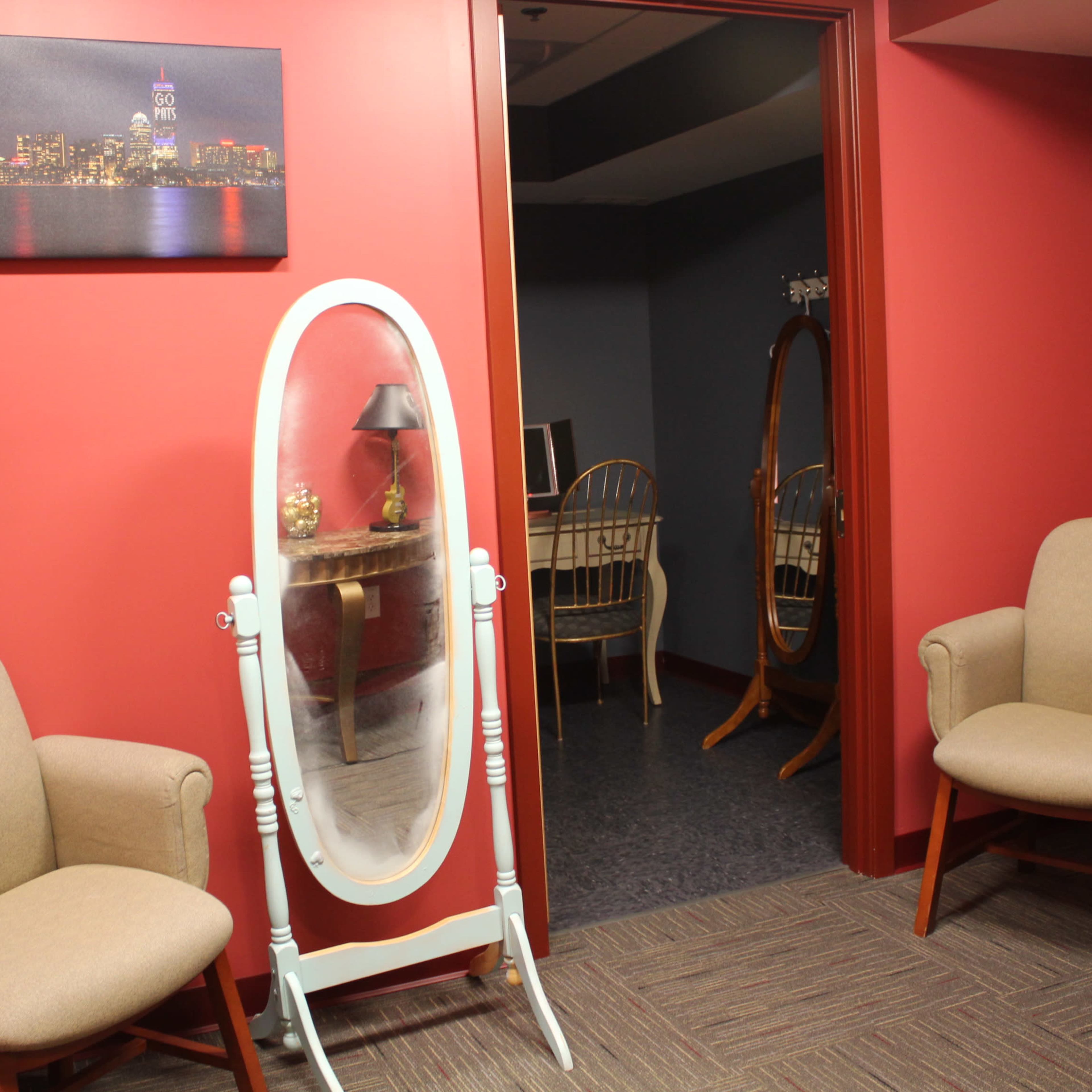 A small waiting area with two beige chairs, a white standing mirror, and a doorway leading to a room with additional furniture in the background.