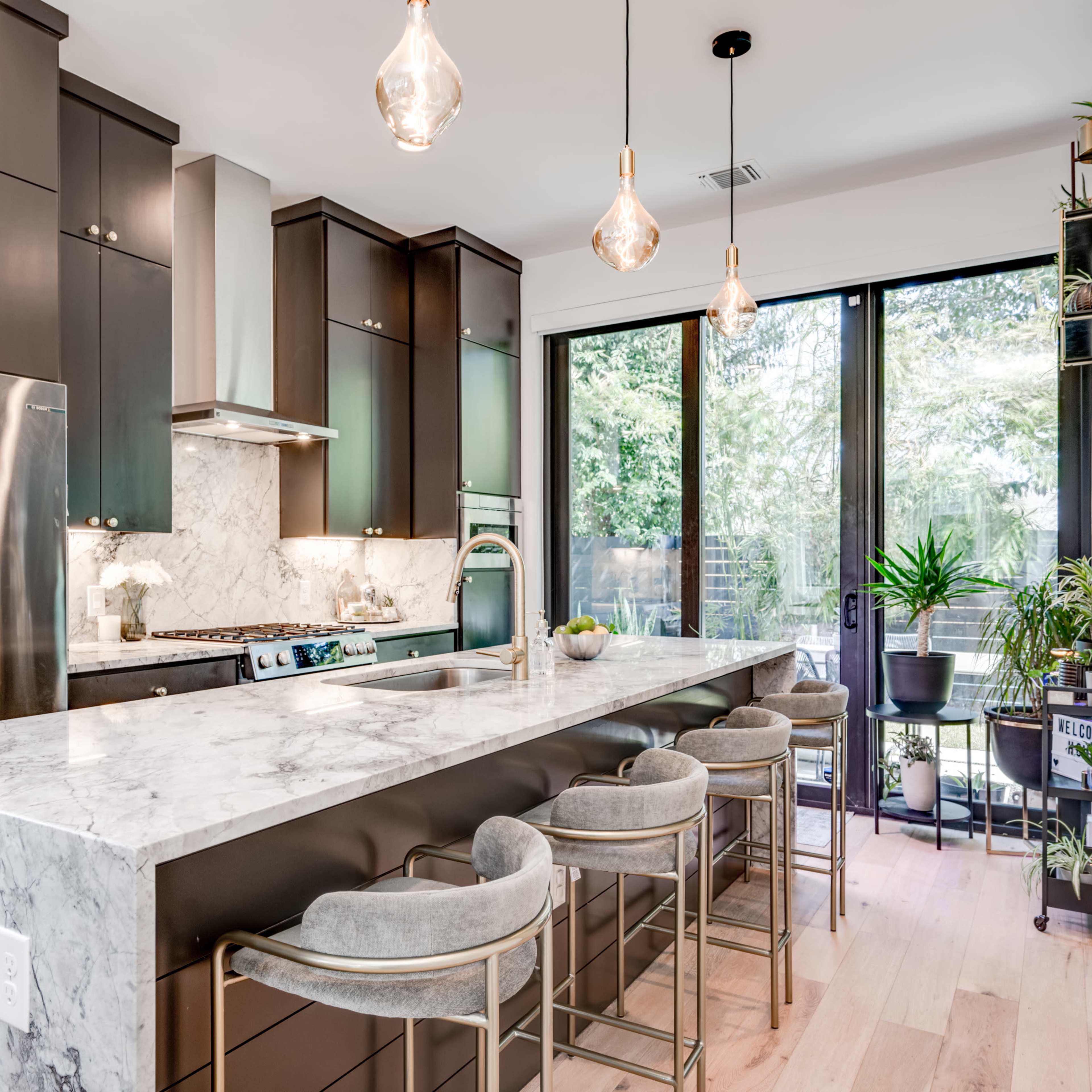 A modern kitchen features dark cabinetry, a marble island, and pendant lighting, with large windows overlooking a patio filled with plants.